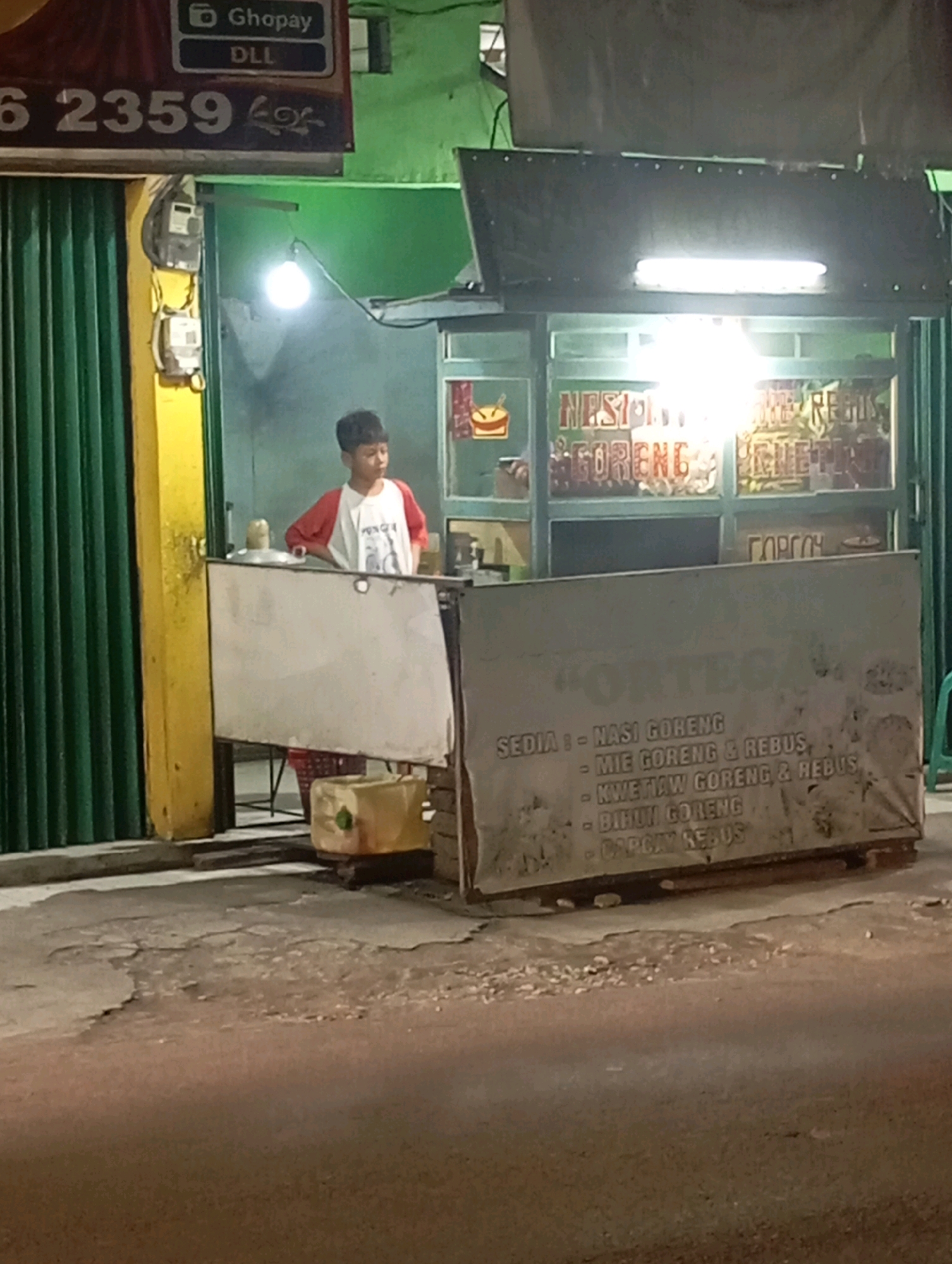 A street food stall operates at night in Babelan, Indonesia. A young boy, wearing a white t-shirt with red sleeves, stands behind the counter. The stall has a sign with the text "Ghopay DLL" at the top, followed by "6 2359" and an ornate symbol. The stall itself is illuminated by fluorescent lights, and a sign above the cooking area displays food items like "NASI GORENG," "MIE GORENG," and "BIHUN GORENG." A grey cloth banner in front of the stall lists menu items: "SEDIA : - NASI GORENG", "- MIE GORENG & REBUS", "- KWETIAW GORENG & REBUS", "- BIHUN GORENG", "- CAPCAY REBUS". The stall is situated on a paved road with visible cracks and debris. A green corrugated metal shutter is visible on the left side of the image.