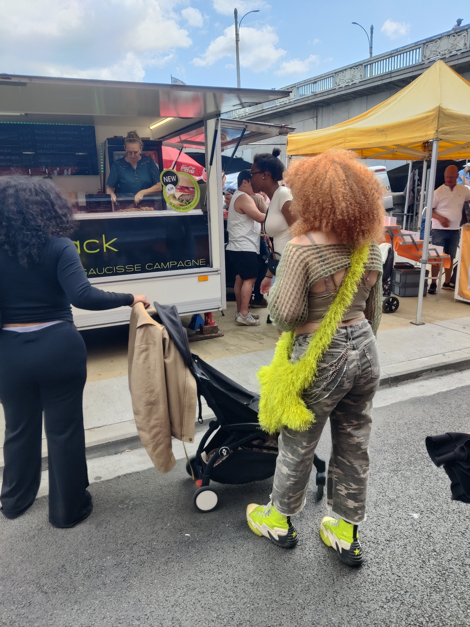 The image depicts an outdoor scene, likely a market or street food event, with a food truck visible on the left and a yellow tent on the right. The food truck has a menu board displaying items such as "SAUCISSE CAMPAGNE" and "BURGER MAISON". A woman in a green shirt is serving customers from the truck. In the foreground, a woman with curly red hair is seen from behind, wearing a green cropped sweater, camouflage pants, and brightly colored sneakers. She is carrying a fluffy yellow bag. Next to her, another woman with dark curly hair is also seen from behind, wearing black pants and a black long-sleeved top. She is holding onto a stroller, which has a tan jacket draped over it. In the background, a concrete bridge with decorative railings spans across the scene, suggesting an urban environment. The presence of food trucks and tents, combined with the style of architecture in the background, is consistent with the atmosphere of the Hors-Château area in Liège, Belgium, known for its vibrant markets and street life. The overall impression is one of a lively public gathering.
