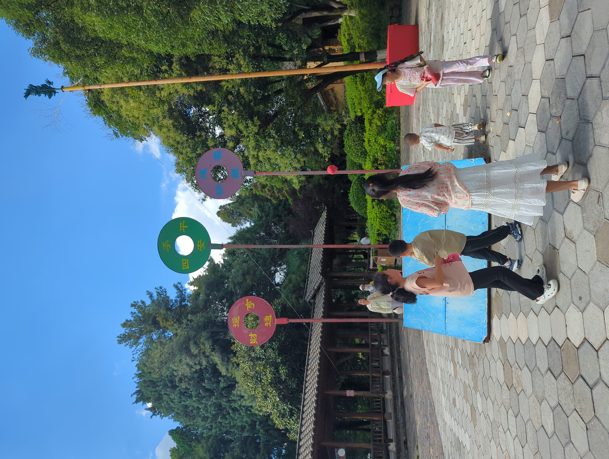 This is a forensic, matter-of-fact description of an outdoor scene in Xishan District, China. The image depicts a bright, sunny day with a clear blue sky and scattered clouds. The foreground is a paved area with a hexagonal stone pattern. Several children are present. Two girls and one boy are standing near a blue structure. One girl, wearing a light-colored dress and white shoes, faces away from the camera and is partially obscuring a boy in a tan shirt and black pants. Another girl, in a white skirt and top, stands to the right, facing left, wearing a blue cap and holding a red object. A young child in a patterned shirt and striped pants walks away from the camera.
In the mid-ground, there are three large, circular objects with Chinese characters, mounted on poles. These objects appear to be decorative elements or part of a game. The leftmost circle is red with yellow characters, the middle is green with yellow characters, and the rightmost is purple with yellow characters. Behind these structures, there is a traditional-looking covered walkway with a tiled roof. Several adults are visible under the walkway.
The background consists of lush green trees and foliage, suggesting a park or garden setting. A tall, wooden pole with a dark, abstract sculpture at the top stands to the right of the frame. To the far right, a bright red rectangular structure is visible. The overall environment appears to be a public outdoor recreational space.