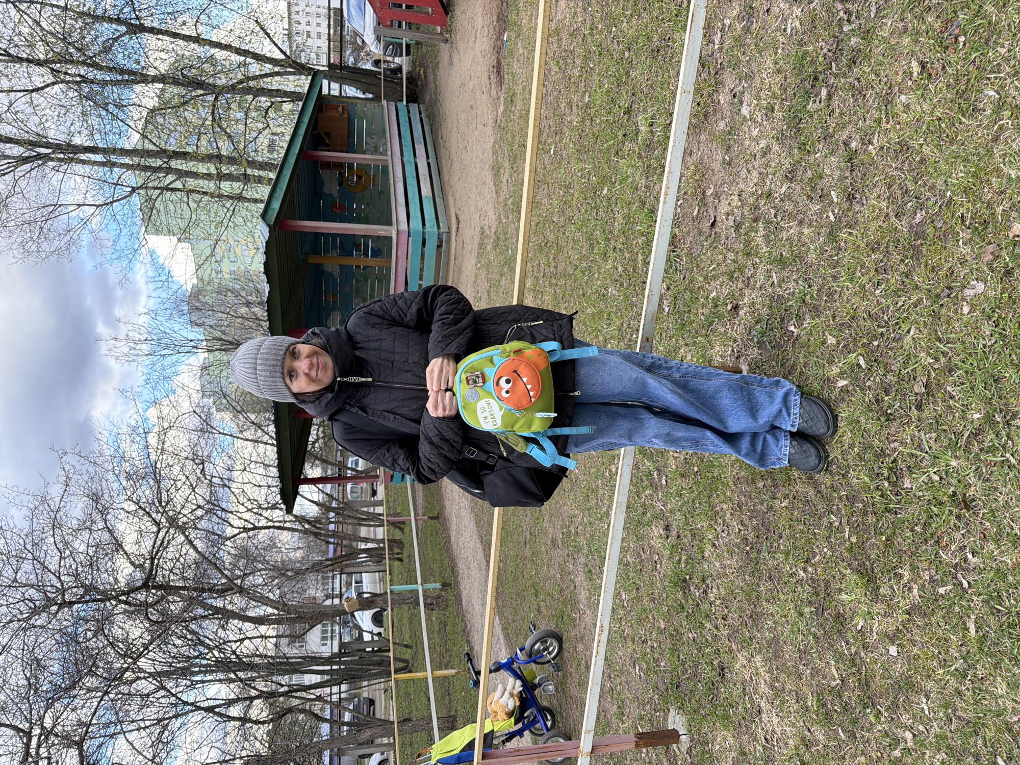The image features a female individual standing outdoors in what appears to be a park or playground in Chaikovsky, Russia. She is smiling faintly at the camera, dressed in a grey knitted beanie, a black quilted jacket, blue jeans, and dark, possibly quilted, flat shoes. In her hands, she holds a small, brightly colored green and orange backpack adorned with a cartoon monster face and the visible text "I'M SO ROARSOME!". She also carries a black shoulder bag on her left side.
The setting is characterized by a mix of sparse green grass and dirt paths, bordered by light-colored metal or wooden rails. Bare deciduous trees with intricate branch patterns dominate the background, suggesting late winter or early spring. Further into the scene, a blue and yellow child's tricycle lies on its side, and a colorful wooden playhouse or kiosk, painted green, blue, and red, is visible. Beyond the trees, several multi-story residential buildings can be seen under a partly cloudy sky with patches of blue. The scene depicts a casual daytime outing, with the individual appearing content and relaxed in the cool, bright weather.