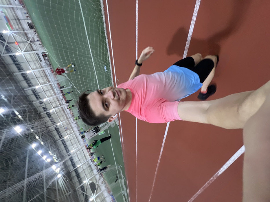 The image captures a selfie taken by a man within an indoor sports facility, likely during or after a sports activity, with a soccer game or practice unfolding in the background. The image is rotated 90 degrees clockwise.
In the foreground, a young man is taking a selfie, holding the camera with his right arm extended. He is positioned on a vibrant red running track marked with white lines. He wears a short-sleeved athletic shirt that features an ombre or gradient effect, transitioning from a bright pink at the shoulders and upper chest to a light blue at the waist. He also wears black athletic shorts and black athletic shoes. A black smartwatch is visible on his left wrist, and white earbuds are in his ears. He has short, dark hair and looks directly at the camera with a relaxed, slight smile. A shadow of his upper body and head is cast on the red track to his right.
The background reveals a large indoor sports area. Behind the man, a green artificial turf field, likely for soccer or futsal, is visible. A white soccer goal net stretches horizontally across the middle ground. Through the net, several other individuals are seen engaged in what appears to be a soccer game or practice. Some players are wearing red jerseys, while others are in green or blue/yellow attire. One player in a red jersey is particularly noticeable near the goal, seemingly in action. The facility features a high ceiling with exposed metal trusses and numerous bright artificial lights, indicating an indoor setting. The overall lighting is strong and even, illuminating the entire space.
