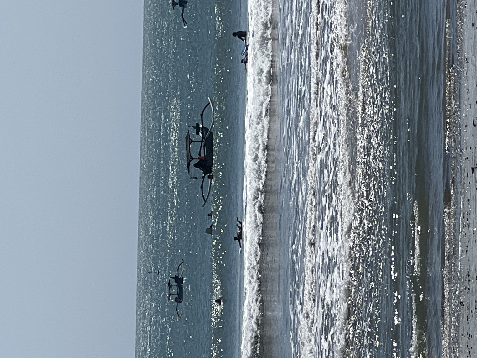 The image captures a serene scene of the ocean at Kuta, Indonesia. Several traditional Balinese fishing boats, known as jukungs, are scattered across the shimmering water. Some of these boats are equipped with canopies, suggesting they are either used for leisure or for transporting people. In the foreground, the waves are breaking, creating a dynamic visual contrast with the calmer waters further out. A few individuals are visible in the water, some appearing to be surfers on their boards, while others are simply enjoying the ocean. The overall atmosphere is one of peaceful coexistence between human activity and the natural beauty of the sea. The sky is clear and bright, indicating a sunny day.