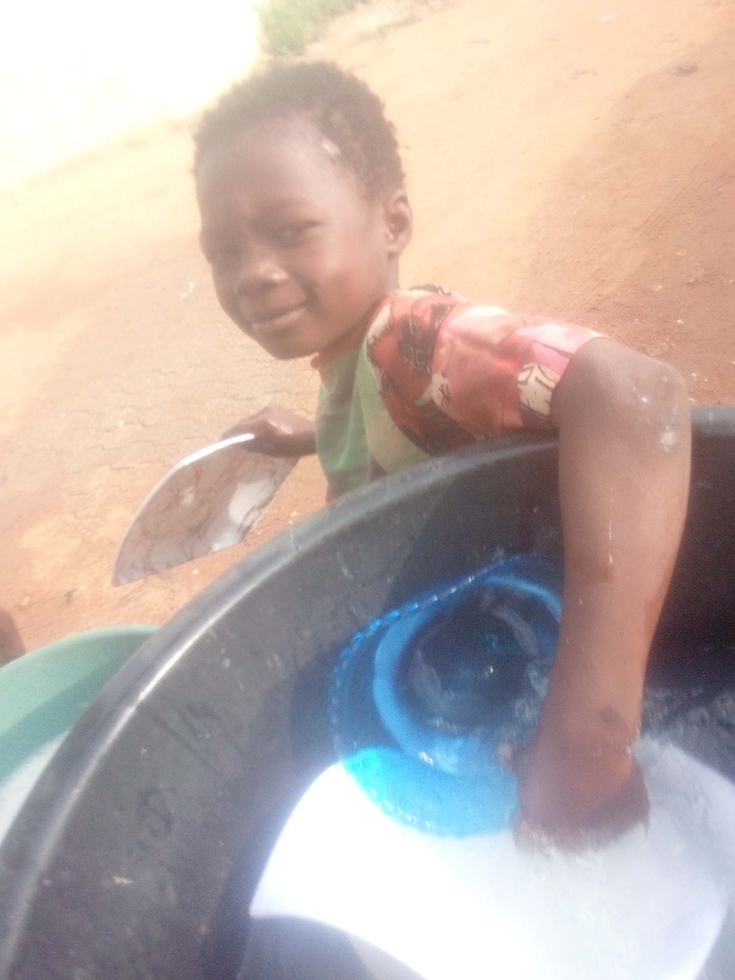 A young child is captured in a close-up, outdoor shot, seemingly engaged in washing. The child, with dark skin and short, dark hair, is looking directly at the camera with a gentle expression. They are wearing a short-sleeved top with a colorful, possibly patterned, design on the shoulders and arms, and a plain green body.
The primary activity taking place is washing, likely dishes or laundry. The child's left hand holds a white plate with subtle dark patterns. Their right arm is immersed in a large, dark-colored plastic basin or tub, which contains water and other items. Within this basin, a blue plastic object, possibly another bowl or container, is partially submerged. The child's right hand appears to be scrubbing or holding a piece of white fabric or clothing in the soapy water. To the left, another light green basin is partially visible.
The setting appears to be outdoors, indicated by the light brown, dusty or dirt ground visible in the background and beneath the basins. The lighting suggests a bright, sunny day, with some overexposure evident in the upper left portion of the image. There are no clear identifying landmarks or visible text in the scene. The child's demeanor seems calm and focused on their task, while also acknowledging the camera.