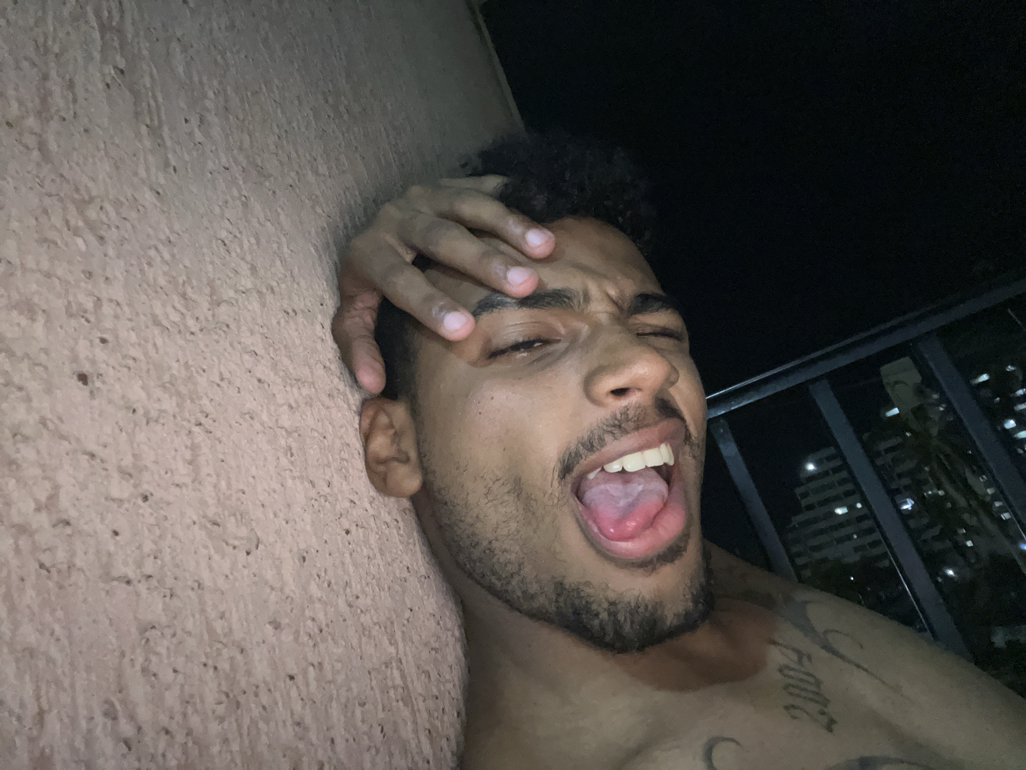 The image captures a young adult male in an outdoor nocturnal setting, identified as the city of Porlamar, Venezuela. The subject is positioned at an angle, leaning or resting against a textured, light-colored wall. He exhibits a dramatic facial expression with squinted eyes and a wide-open mouth, revealing his tongue and upper teeth. His right hand is placed on his forehead, partially obscuring the upper part of his face. The male subject has dark, curly hair and a beard/stubble. He is shirtless, displaying a tattoo of the numerical sequence "2004" on his right shoulder/upper arm area, and another partial tattoo beneath it. To the right of the subject, a dark background reveals a black metal railing and indistinct city buildings with illuminated windows in the distance, indicative of nighttime urban environment. The scene is illuminated by an unseen light source that highlights the subject and the immediate wall, contrasting with the darker, ambiently lit background.