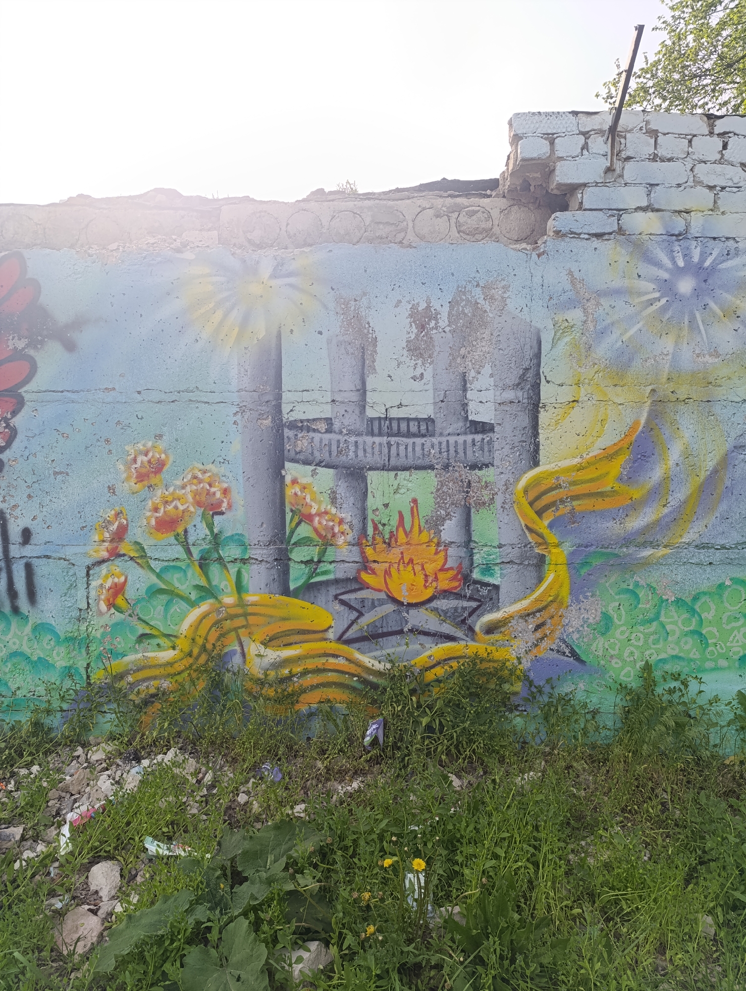 An eye-level outdoor shot shows a graffiti mural on a deteriorating wall in an unknown city location. The mural depicts an industrial scene with smokestacks, flowers, a starburst-like shape, and a yellow ribbon-like element. The wall is light blue and green, with a section of exposed brick at the top right. The foreground is overgrown with weeds and grass. The scene conveys a sense of decay and neglect.