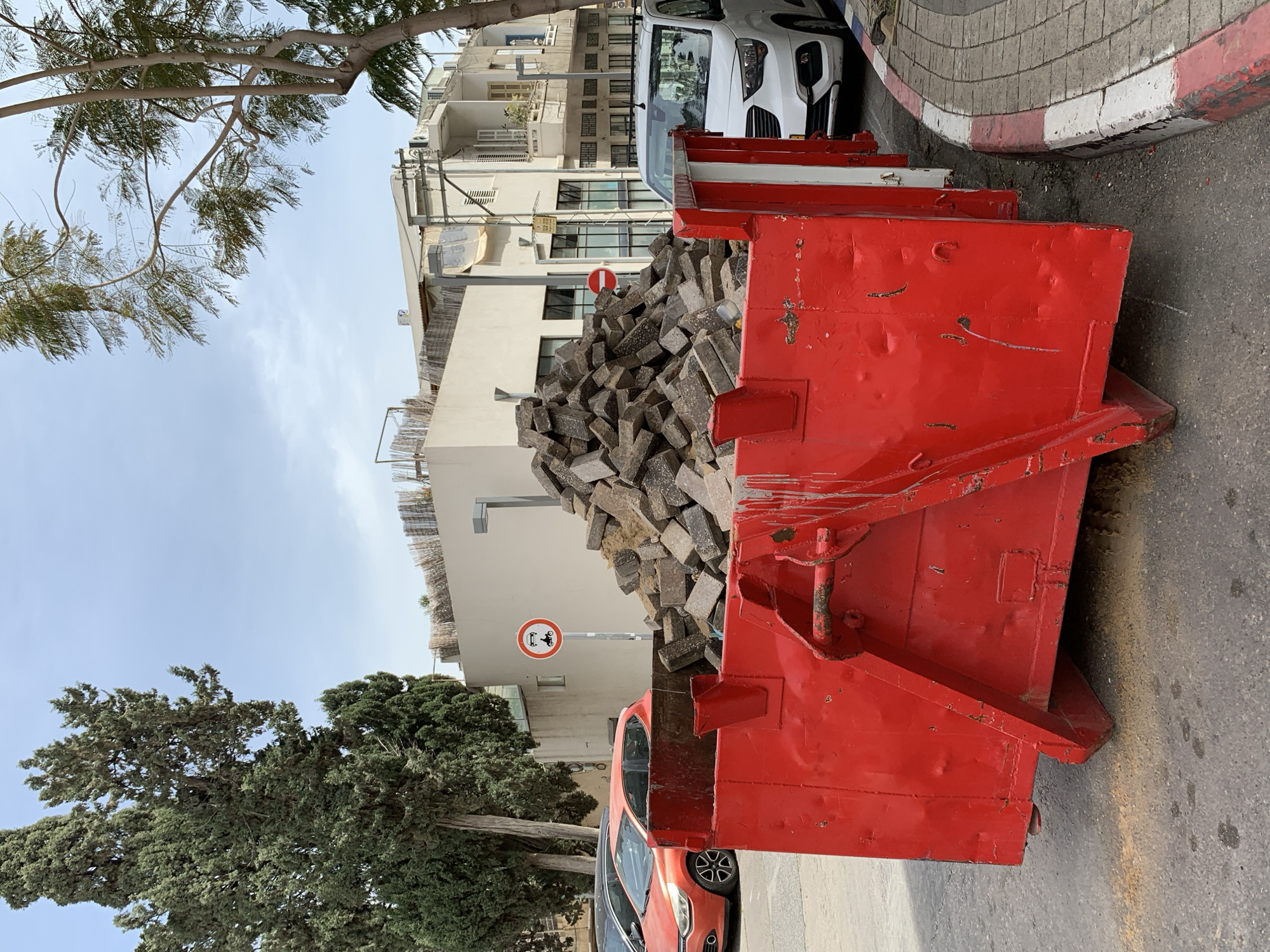 The image shows a red construction dumpster overflowing with gray bricks, parked on the side of a street in Tel Aviv, Israel. The curb is marked with a red and white stripe. A white van is parked behind the dumpster, and an orange car is visible on the other side. A "No Parking" sign with a motorcycle symbol is nearby. Buildings with balconies and a tree with sparse leaves are in the background, with blue sky above.