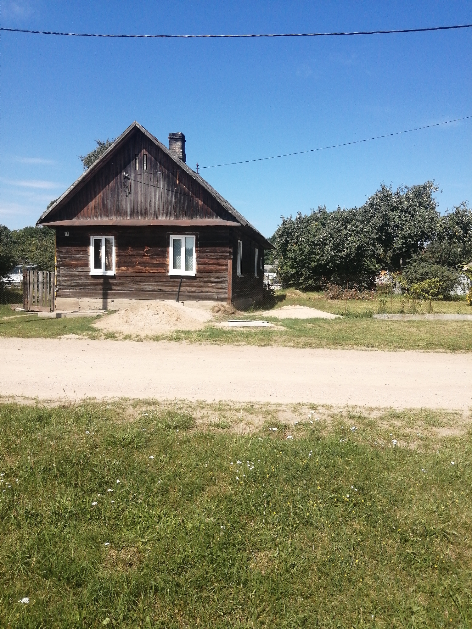 The image depicts a single-story wooden house with a gabled roof and a brick chimney, situated in the city of Slonim, Belarus. The house has white-framed windows. In front of the house, there is a pile of sand, a wooden gate, and a dirt road. The area surrounding the house is covered with green grass and scattered small white flowers. Lush green trees are visible in the background. The sky is clear and blue. There are no people visible in the image. The scene appears to be a rural or semi-rural residential area.