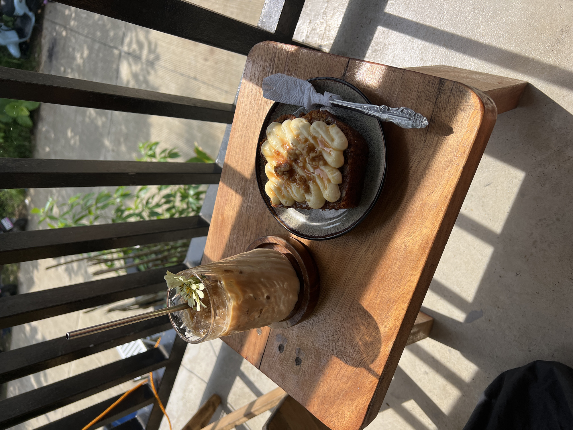 A square-shaped slice of cake with cream cheese frosting and walnuts, drizzled with caramel sauce, sits on a plate next to a glass of iced coffee topped with a flower and a metal straw. Both are placed on a wooden table with a metal railing in the background. The scene appears to be outdoors, possibly a cafe balcony, in San Isidro, Philippines, as indicated by the sunlight casting shadows and the lush greenery visible behind the railing.
