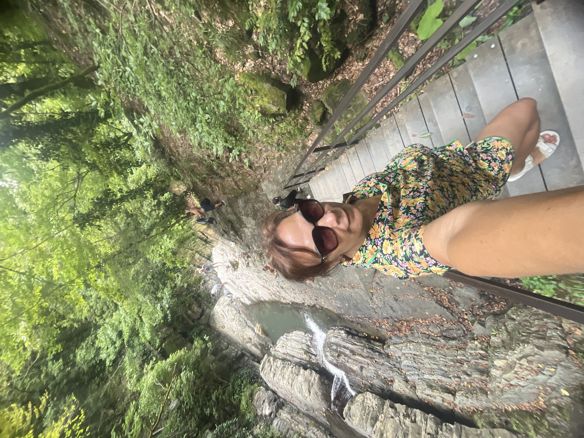 The image shows a woman taking a selfie on a wooden staircase in a lush green forest. She is wearing a floral dress and sunglasses. Behind her, a waterfall cascades down a rocky ravine. Other people are visible in the background, hiking down the path. The scene appears to be in the mountains near Khostinskiy, Russia, a region known for its natural beauty and waterfalls. The overall atmosphere is one of outdoor recreation and enjoying nature.