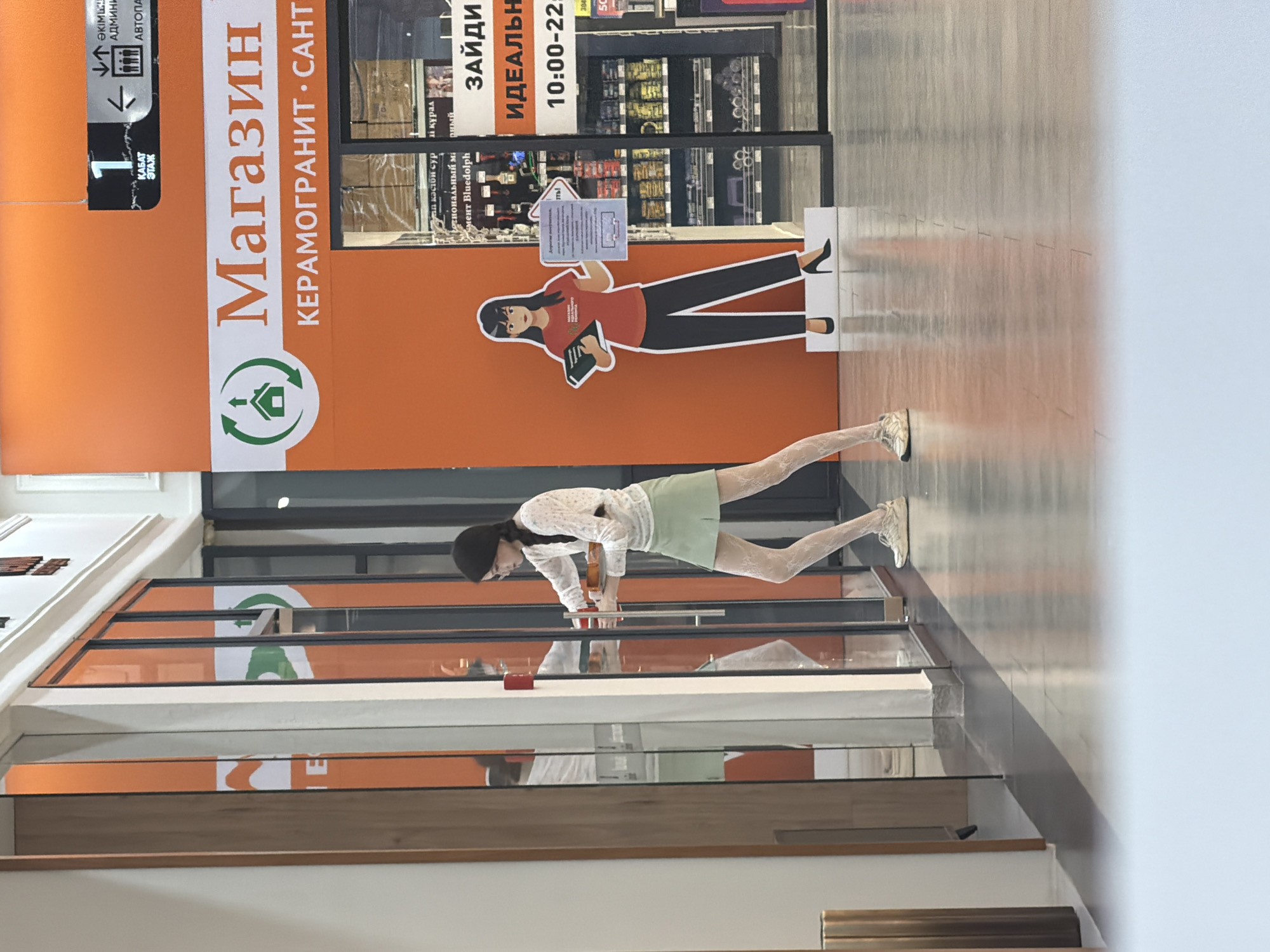 The image depicts the interior of a modern shopping mall in Astana, Kazakhstan, specifically in front of a store entrance. The store appears to be a retailer of ceramic tiles and sanitary ware, as indicated by the large sign displaying "Магазин" (Shop) and "КЕРАМОГРАНИТ • САНТ" (Ceramic Granite • Sant). The store's interior is visible through glass doors, revealing shelves stocked with various products.
In the foreground, a young woman with dark hair, styled in a braid, is seen interacting with the store's automatic sliding glass doors. She is wearing a white, long-sleeved blouse with a floral pattern, a light green pleated skirt, and white lace-patterned tights. Her feet are clad in athletic shoes. She is holding a tray with items that appear to be food or beverages.
To the right of the store entrance, a life-sized cardboard cutout of a woman in a red t-shirt and black pants stands, holding a book and a sign. The sign, written in Russian, appears to be informational. Another sign above the store entrance indicates "1 КАБАТ ЭТАЖ" (1st Floor), along with directional arrows for "АДМИНИ" (Administration) and "АВТОПА" (Autopark).
The mall's flooring is a polished grey material, reflecting the overhead lighting and the vibrant orange wall behind the store. The overall environment is clean, well-lit, and suggests a commercial setting. The reflection in the glass doors shows another person inside the store, partially obscured. The lighting casts distinct shadows on the floor, indicating the direction of the light source.