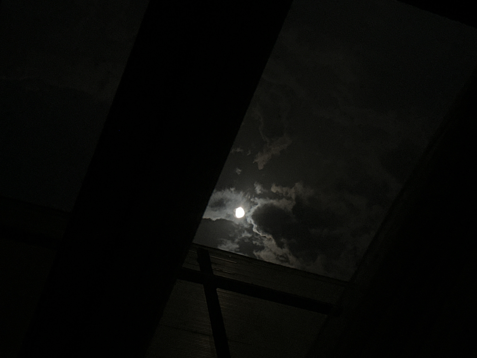 The image depicts a view of the night sky from indoors, likely taken in Yerevan, Armenia. A bright moon is visible through a rectangular opening, obscured partially by clouds. The dark shapes of interior structures, possibly a window frame or beams, frame the view of the sky. The scene is generally dark, with the moon being the primary light source.