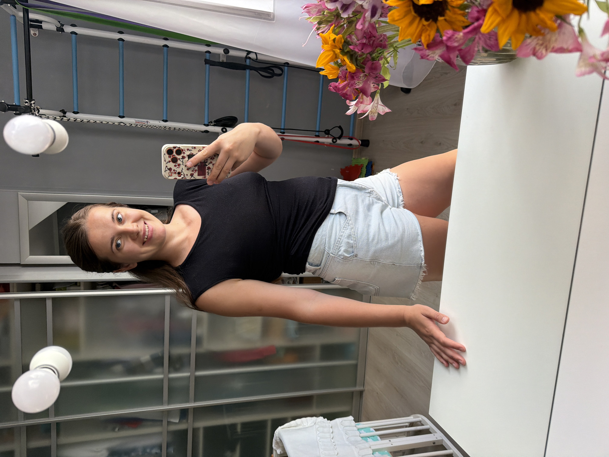A young woman with brown hair pulled back takes a selfie in a brightly lit room, possibly a bedroom or home gym. She is wearing a black sleeveless top and light blue denim shorts with a frayed hem. Her left hand rests on a white table in the foreground. In the reflection of the mirror, a large closet with mirrored doors is visible behind her. To her right, there is a wall-mounted exercise ladder with blue rungs and various straps hanging from it. A bouquet of colorful flowers, including sunflowers, is arranged in a glass vase on the table to the right of the frame. A white crib or bassinet is partially visible on the left side of the image. The flooring appears to be light-colored wood laminate. There is no indication of the city of Sochi, Russia, in the image itself.