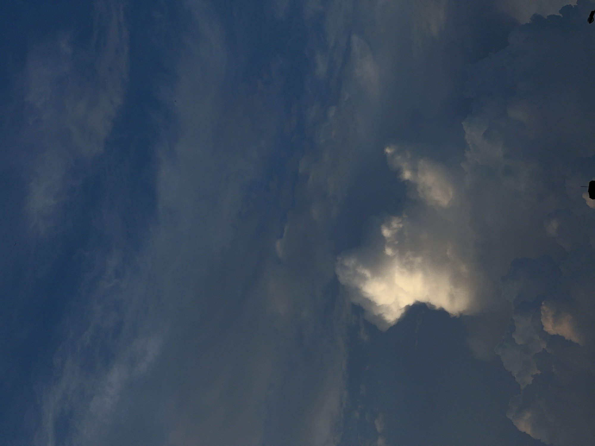 The image shows a dark blue sky with various clouds. In the center of the image, a bright white cloud stands out against the darker sky. The bottom of the image shows the silhouettes of buildings and a lamppost against the sky. There are no people or animals visible in the image. The scene appears to be a view of the sky from a city. The location is Krasnodar, Russia.
