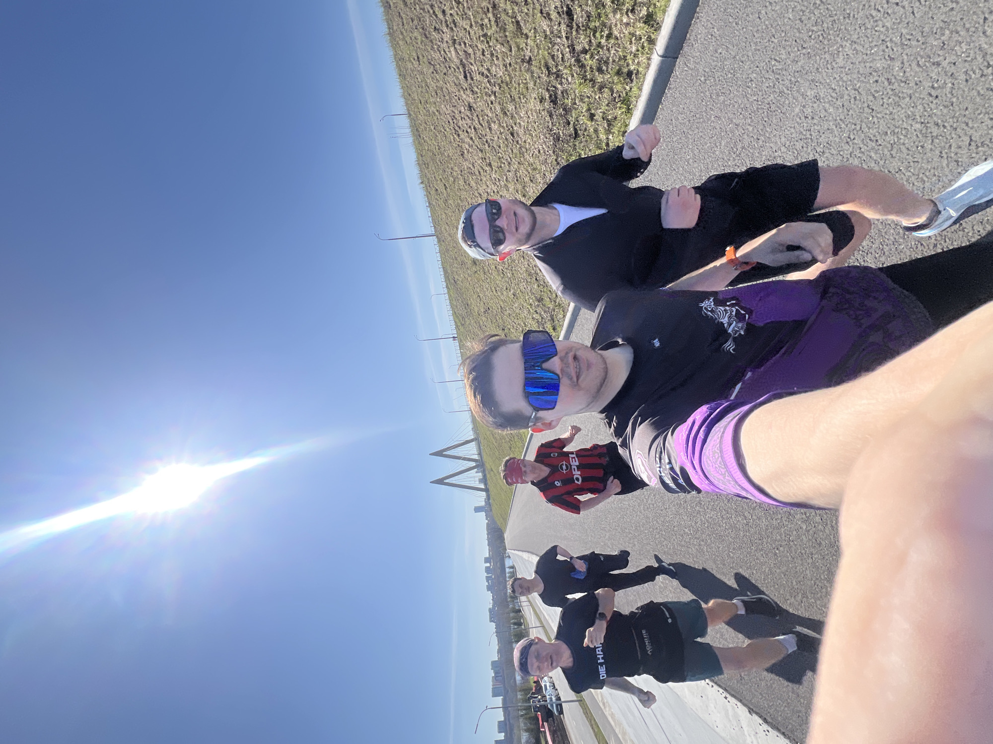 The image is a selfie taken on a clear, sunny day with a bright sun in the sky, likely in Kazan, Russia, as evidenced by the prominent cable-stayed bridge and urban landscape.
Two men are positioned closest to the camera, one wearing blue-tinted sunglasses, a black shirt with a purple design and purple shorts, and the other wearing sunglasses, a black jacket over a white shirt, and black shorts. The man closer to the camera appears to be holding the camera to take the selfie.
Behind them, several other individuals are present. One wears a red and black striped soccer jersey with a red bandana, and a couple of others are further back, dressed in black running attire. These individuals appear to be running along the paved area.
The setting appears to be a walkway or path alongside a road, and behind them is a grassy area leading towards the bridge and the city skyline. The bridge is a notable landmark suggesting the location is Kazan, Russia.