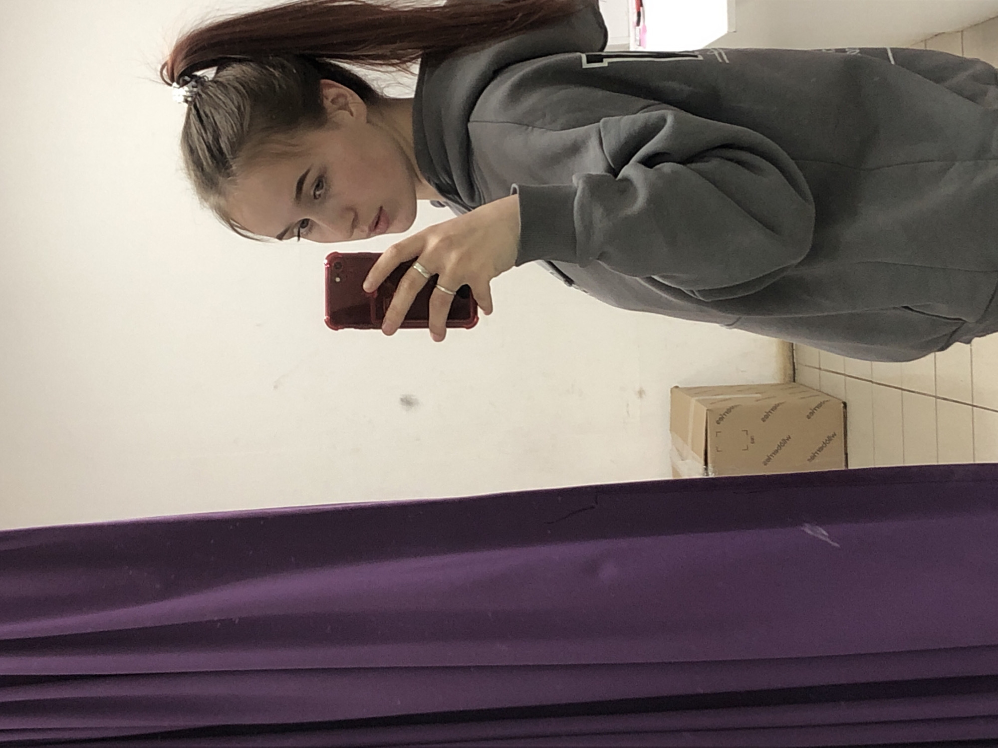 The scene depicts a young female inside what appears to be a changing room or retail space in the city of Фоки, Russia. The female, with brown hair tied in a high ponytail secured by a metallic-colored scrunchie, wears a grey hooded sweatshirt with white markings on the right side. She is holding a dark red smartphone with a matching protective case in her left hand, positioned for a mirror selfie. Two silver-colored rings are visible on the fourth and fifth fingers of her left hand. She is looking slightly to her left, past the camera. The background consists of a plain off-white wall on the right and a dark purple curtain on the left, which has minor scuffs and marks. On the floor, which is tiled with light-colored squares and dark grout lines, a closed, light brown cardboard box with reversed text is visible. A small white surface is partially visible on the far right.