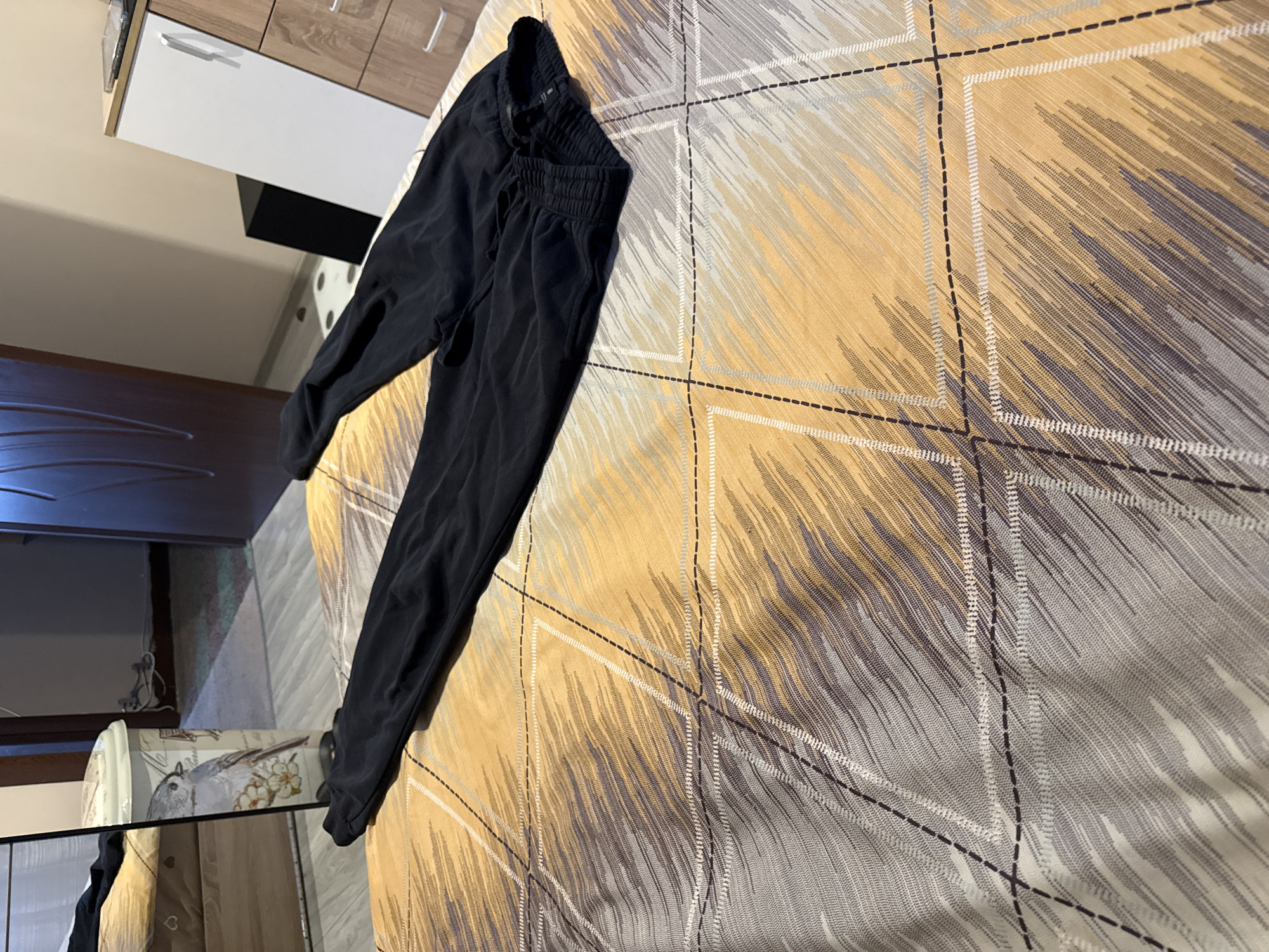 The image shows a pair of black sweatpants lying on a bed with a patterned bedspread. The bedspread features an abstract design in shades of yellow, grey, and brown, with darker stitched lines forming a grid pattern. To the left of the bed, a partially visible bedside table with white drawers and wooden trim is present. In the background, parts of the room are visible, including what appears to be furniture with a dark wood finish.
While the prompt mentions Lovech, Bulgaria, there is no information in the image to definitively confirm this location.