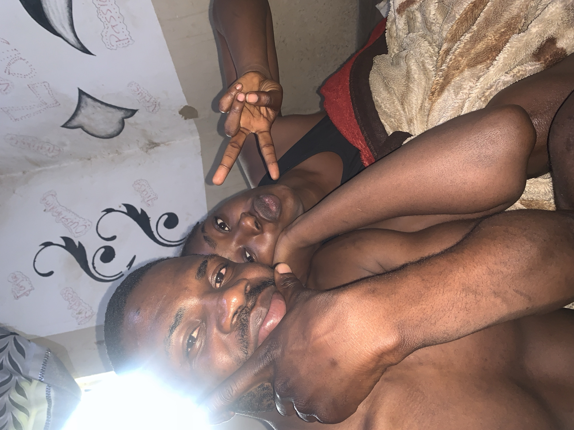 The image is a close-up selfie taken indoors, likely in Rafin Par, Nigeria, showing a man and a woman embracing. The man's face is in the foreground, looking towards the camera, with the woman's face partially visible behind him, resting her head on his shoulder. She is making a peace sign with her right hand. They are both dark-skinned and appear to be young adults. The background shows a wall with some artistic drawings, possibly of flowers and text, and a blanket is visible draped over them. The lighting suggests it is daytime, with a strong light source coming from the left side of the image.