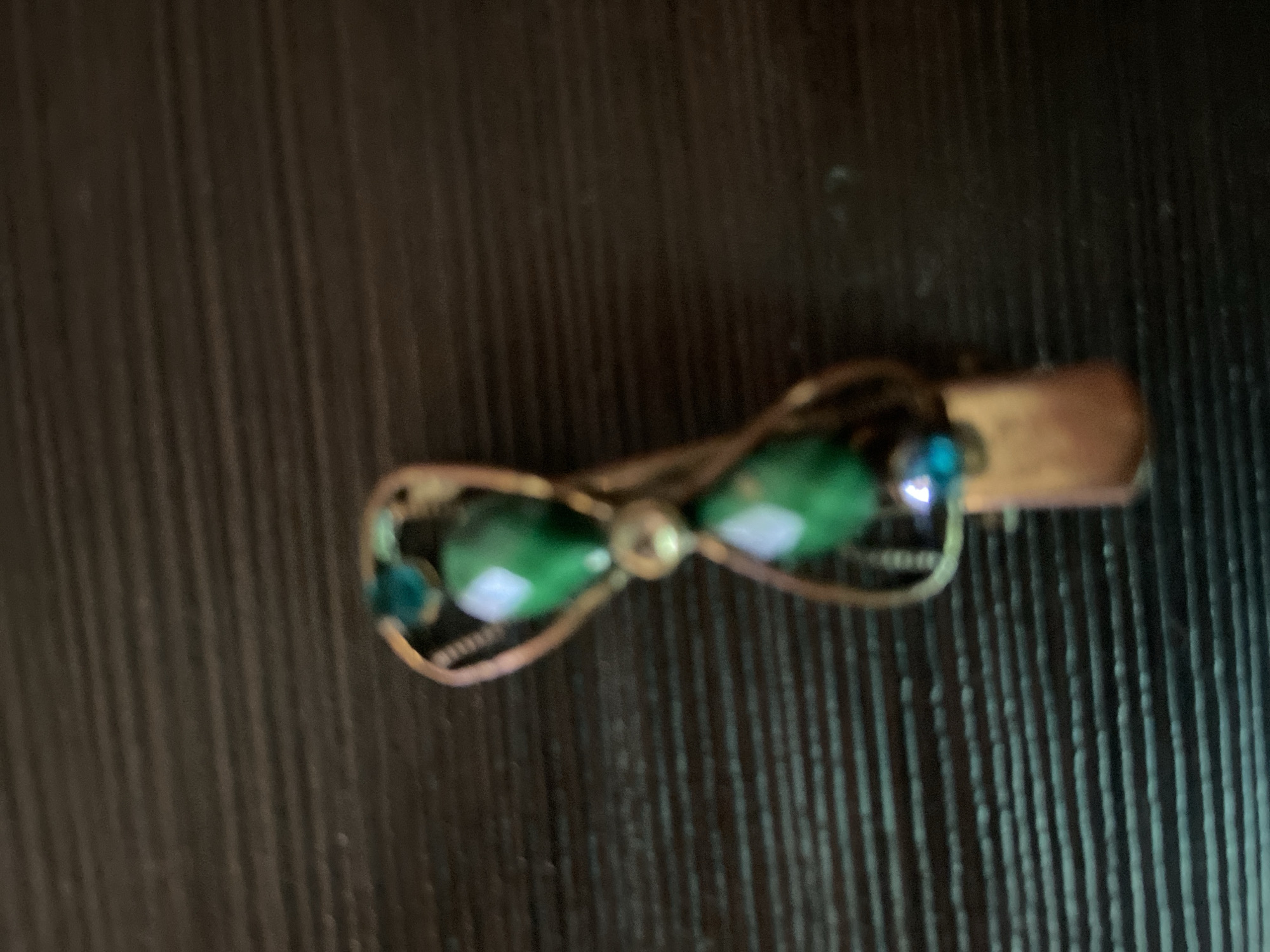 A close-up, slightly blurry, top-down shot shows a hair clip with green, teardrop-shaped beads and a small blue bead, set against a dark, horizontally ribbed surface. The hair clip appears to be made of a brass-colored metal. The background surface has fine parallel lines, creating a textured appearance. The lighting suggests the object is indoors, with no indication of a specific location in a city.