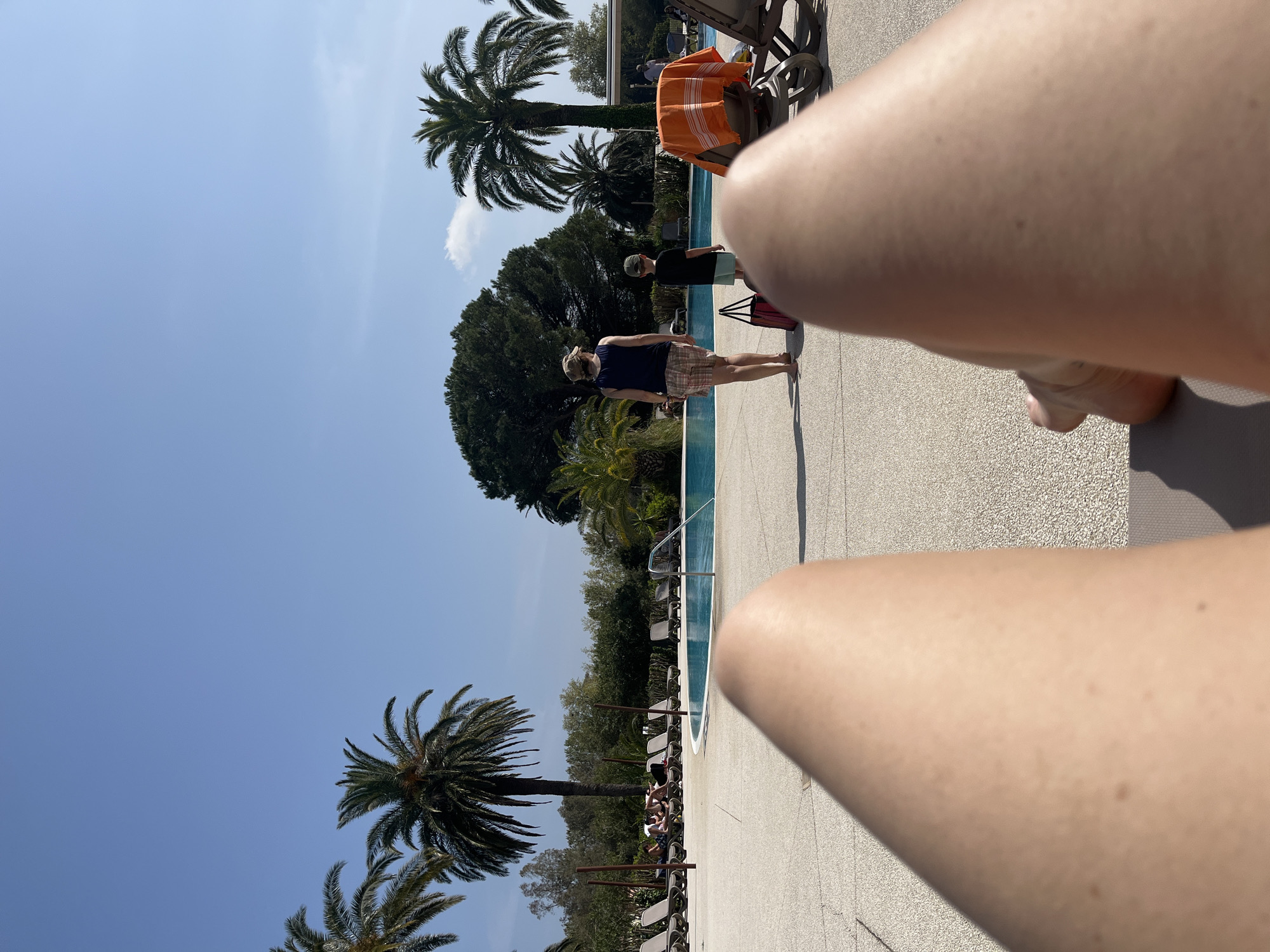 The image, taken from a first-person perspective, shows a poolside scene in Grimaud, France. The foreground is dominated by the photographer's legs, stretched out on a lounge chair. In the background, a swimming pool is visible, bordered by palm trees and other greenery. Two people are walking alongside the pool: one wearing a blue top and plaid shorts, and another in a black shirt and shorts. Lounge chairs are arranged along the poolside, some occupied. An orange towel is draped over one of the chairs near the pool. The sky is clear blue with scattered clouds.