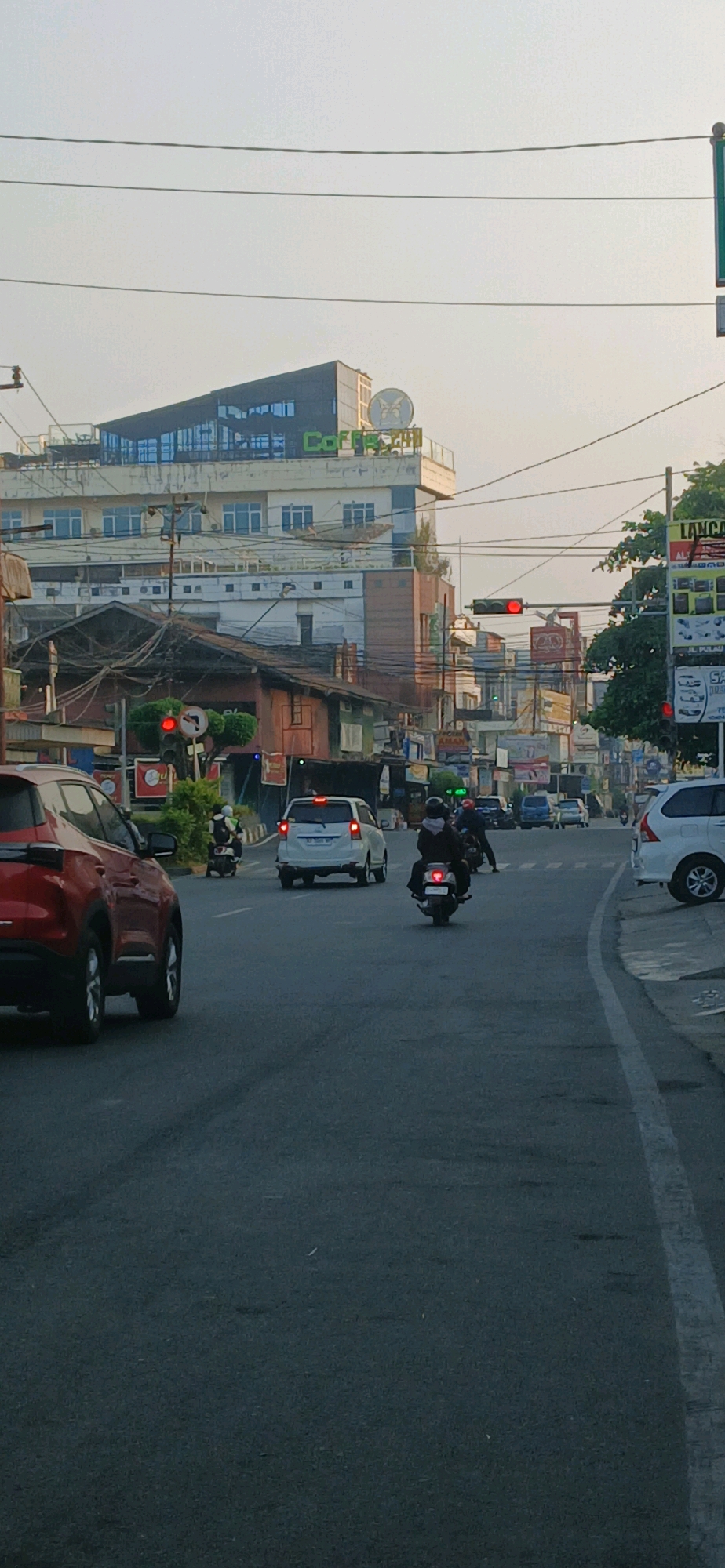 The image depicts a street scene in Samarinda, Indonesia, likely during daylight hours with a bright sky. In the foreground, a red SUV is visible on the left, and a white sedan and a motorcycle are further down the road. Several other vehicles, including cars and motorcycles, are present in the middle ground. A traffic light is illuminated red, indicating a stop.
Buildings line both sides of the street. On the left, a multi-story building with a modern, angular rooftop structure featuring large windows and a sign that appears to say "Coffee Time" is prominent. Other buildings of varying heights and architectural styles are visible behind it. On the right side of the street, there are more commercial buildings with signs, including one that reads "LANCAR." The environment appears to be a busy urban setting with overhead power lines crossing the street. The road surface is asphalt and marked with white lines. The overall impression is one of a typical city street with moderate traffic.