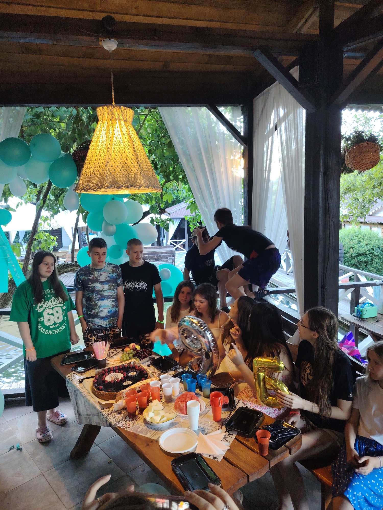 This image captures a birthday celebration outdoors, likely in Rostov-on-Don, Russia, given the context provided. The scene is set under a wooden-beamed canopy, adorned with a decorative woven lampshade, with sheer white curtains framing the open sides. Green foliage and trees are visible in the background, suggesting a park or a backyard setting. The celebration centers around a wooden table laden with food and decorations. A large, dark chocolate cake with berry toppings and the number "6" prominently displayed takes pride of place. Surrounding the cake are various party essentials: colorful paper cups, plates, a bowl of cut fruit, grapes, and what appear to be straws. Several people are gathered around the table, engaged in conversation and celebrating. Among them are young men and women, some dressed casually in t-shirts. The presence of teal and white balloons adds to the festive atmosphere. In the background, a man is seen in a dynamic pose, perhaps jumping or playing, while another person is seated on a railing. The overall impression is one of a lively and joyous birthday party.