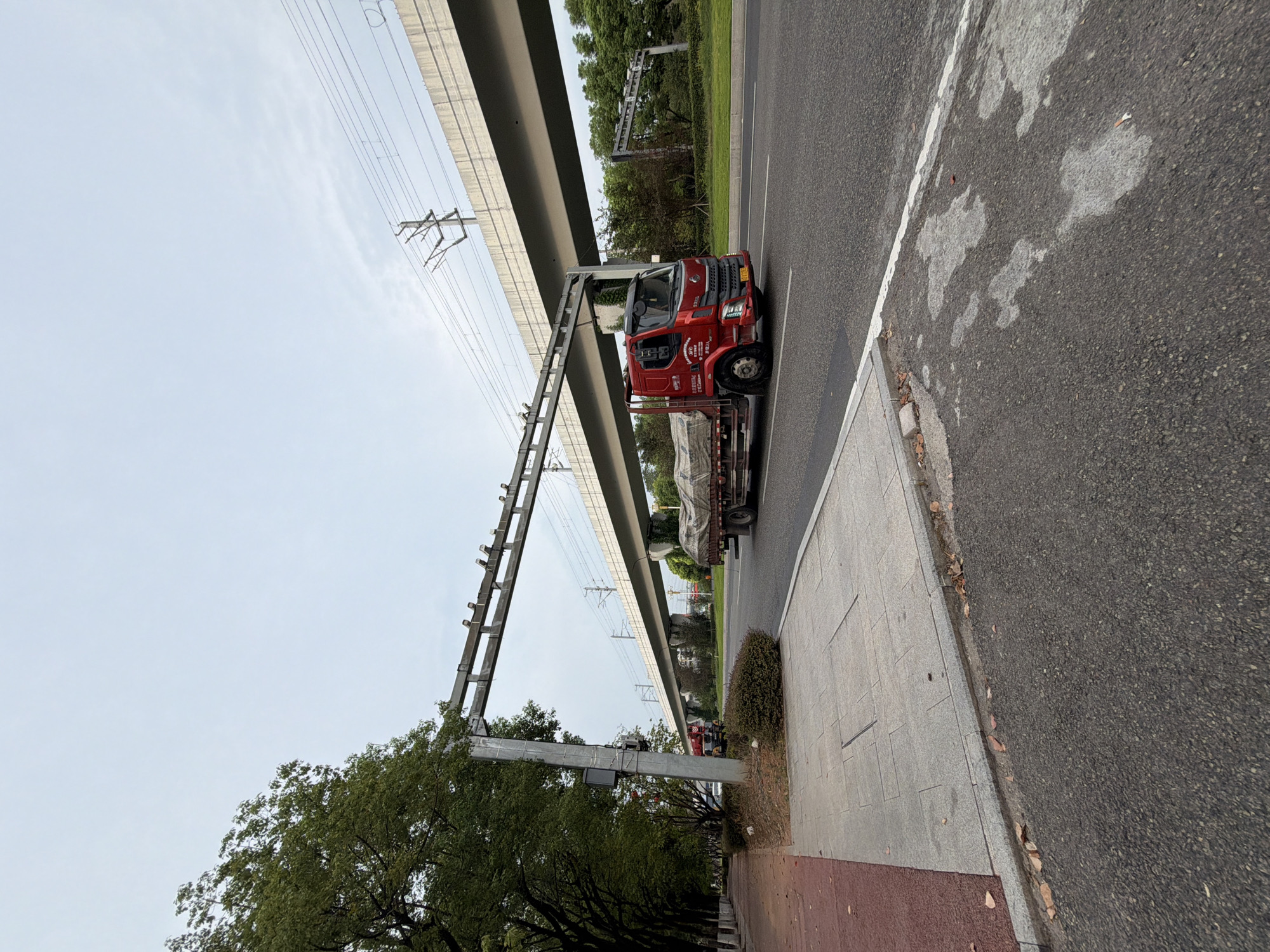 The image depicts a red truck driving on a road in Jiangjin, China. The truck is carrying a load covered by a tarp. In the background, there is an elevated structure, likely a bridge or an overpass, with overhead power lines. A sidewalk runs alongside the road, lined with trees. The sky is overcast. The scene appears to be a typical urban or suburban environment.