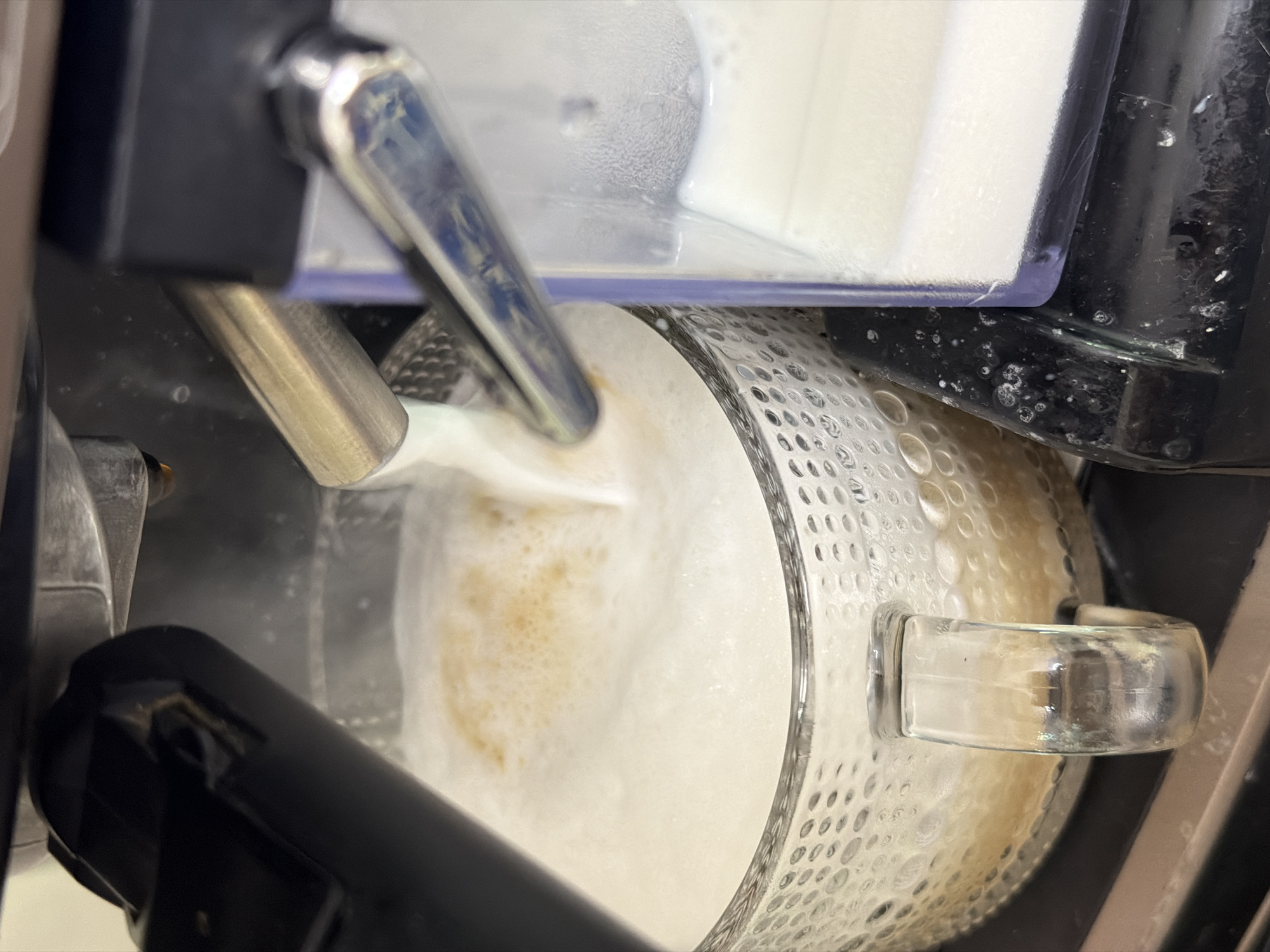 The image depicts a close-up view of a coffee machine dispensing frothed milk into a glass mug. The mug is made of clear glass with a textured pattern and contains a foamy, light brown liquid. A metal spout from the coffee machine is submerged in the liquid, releasing more foam. To the right of the mug is a clear container, possibly for milk, with a white substance inside. The machine itself appears to be made of black plastic and metal components. The overall scene suggests the preparation of a hot beverage, likely a cappuccino or latte. The image does not contain any identifiable landmarks or elements that would indicate its location in the city of Sochi, Russia.