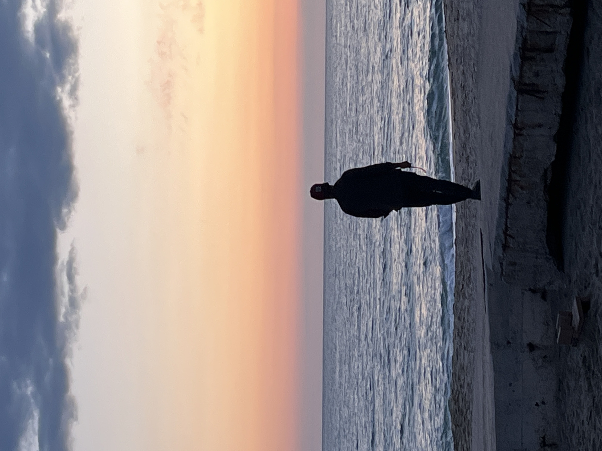 The image depicts a person standing on a beach near the Mediterranean Sea in Alanya, Turkey. The sky is divided between clouds on the left and a pastel sunset on the right. The person, silhouetted and facing right, appears to be wearing a dark outfit and a dark baseball cap with a red top. The sea is visible on the right, with gentle waves approaching the shore. Behind the beach, there's a concrete structure, potentially a seawall or promenade. The photograph has been taken vertically.