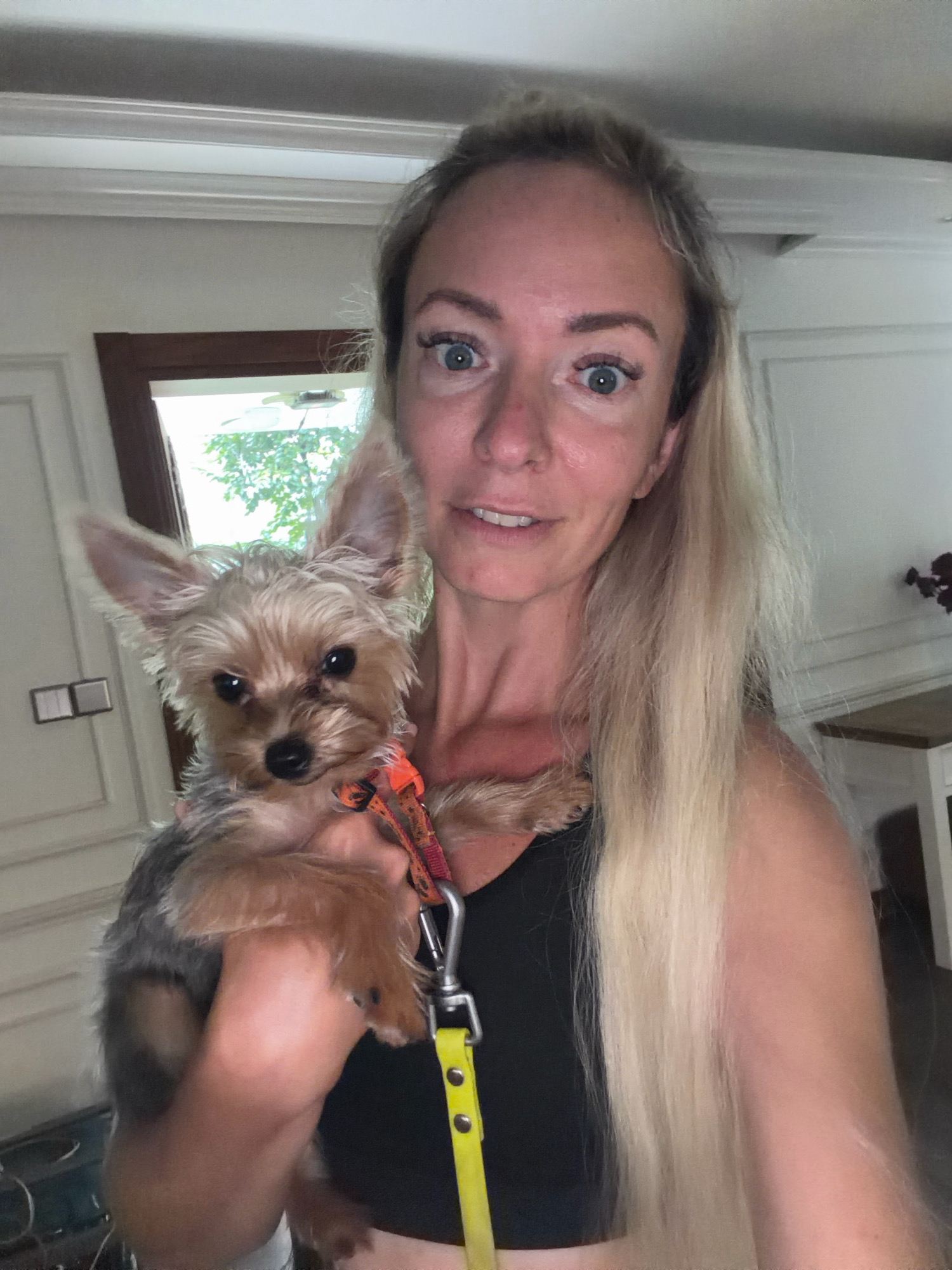A fair-skinned woman with blonde hair is holding a Yorkshire Terrier dog. She is looking at the camera with a surprised expression. The dog is looking to the left, with its ears perked up. Both subjects are wearing collars. The woman is wearing a black tank top, and the dog is wearing an orange collar with a leash attached. The background is blurred, but it appears to be an indoor setting with a window and a door. The location is identified as Xuân Trường - Đà Lạt, Vietnam.