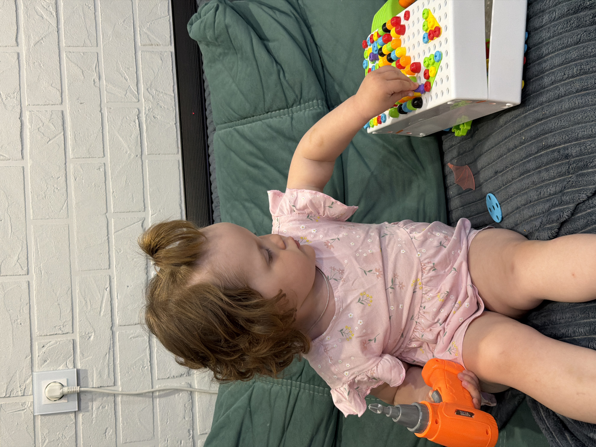 A young child sits on a couch, engrossed in playing with toys. The child wears a light pink floral patterned garment and has brown hair styled in small pigtails. In one hand, the child holds an orange toy drill, while the other hand engages with a white plastic toy with colorful geometric shapes. The toy rests on the couch alongside the child. The couch itself appears to have both a green and a gray section, with varying textures. A white brick wall is visible to the left, featuring an electrical outlet. The photograph provides no data to confirm a link with the city of Pervomaysky, Russia.