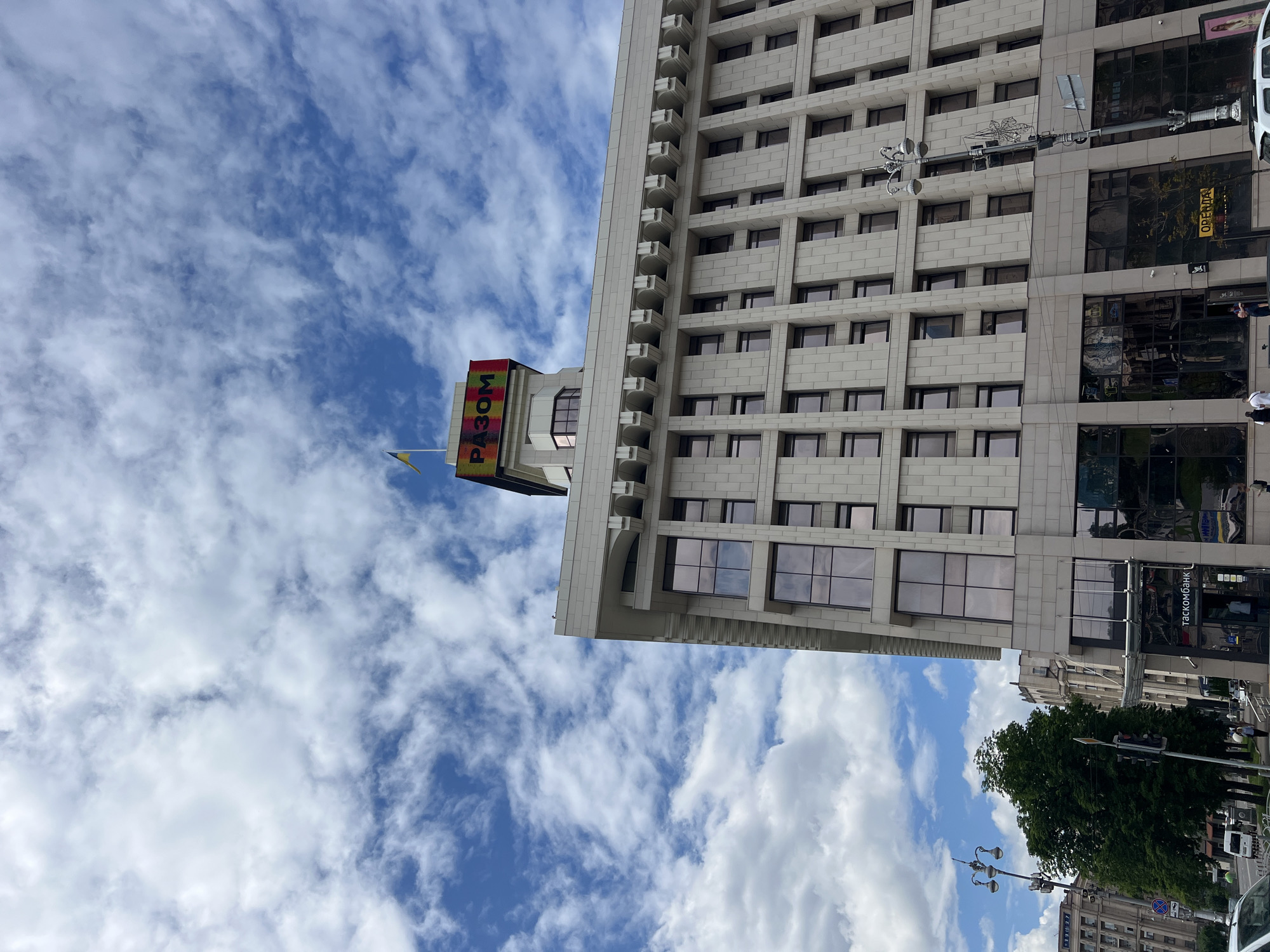 A low-angle medium shot captures a modern, light-colored stone-clad building in Kyiv, Ukraine. The building has a grid of dark-tinted windows and a decorative cornice. Atop the building, a sign with the multicolored Ukrainian word "РАЗОМ" (RAZOM) is visible, topped by the Ukrainian flag. The sky above is bright blue with scattered white clouds. In the lower portion of the image, a street with trees, a lamppost, a traffic light, and pedestrians is partially visible, indicating an urban setting.