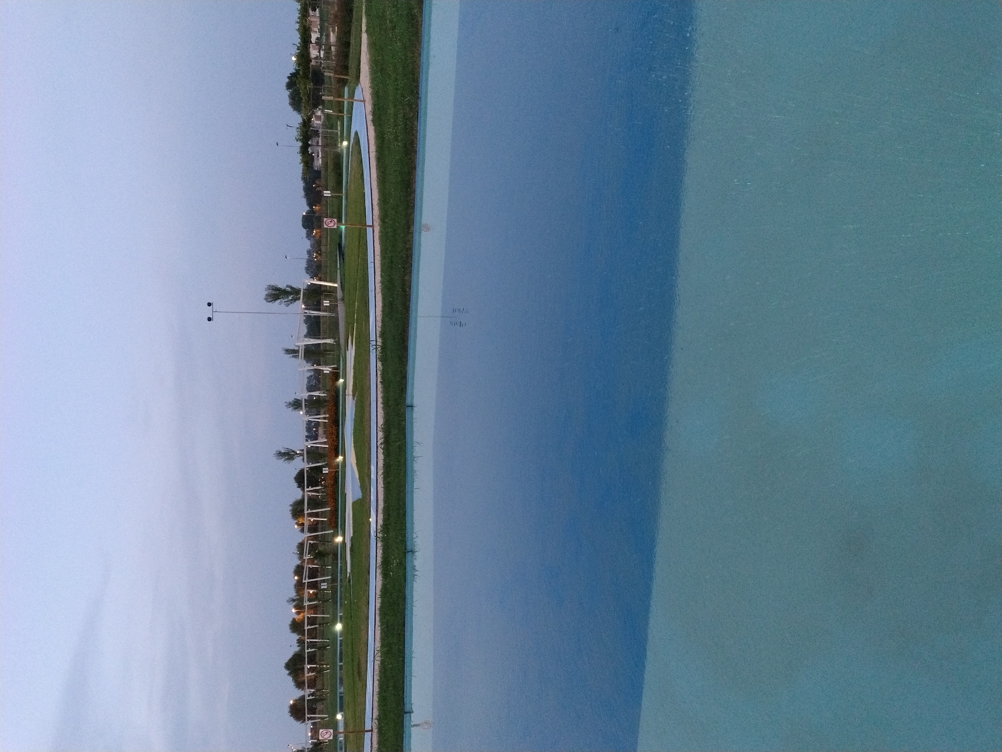 The image depicts a dusk scene in a park in Caorle, Italy, likely during the summer season. In the foreground, a large, shallow body of water with a blue and turquoise gradient occupies the lower half of the frame. The water's surface reflects the sky and hints at submerged features. Beyond the water, a manicured lawn transitions into a paved pathway with a light blue border. This pathway curves through the park, bordered by grassy areas and low-lying bushes.
In the middle ground, several white, arch-like structures are visible, spaced evenly along the pathway. These structures appear to be part of a playground or an aesthetic installation. White streetlights with multiple bulbs are strategically placed throughout the park, illuminating the pathways and green spaces. A few trees with full canopies dot the landscape, and a taller, slender tree stands out on the left. Two circular signs with a red border and a white circle inside, likely indicating rules or restrictions, are visible near the pathway.
In the background, a line of trees forms a backdrop, with more distant structures and streetlights suggesting the edge of the park or a nearby urban area. The sky above is a pale, dusky blue, with subtle cloud formations, indicating the transition from day to night. The overall atmosphere is serene and tranquil, suggesting a public recreational area designed for leisure and relaxation.