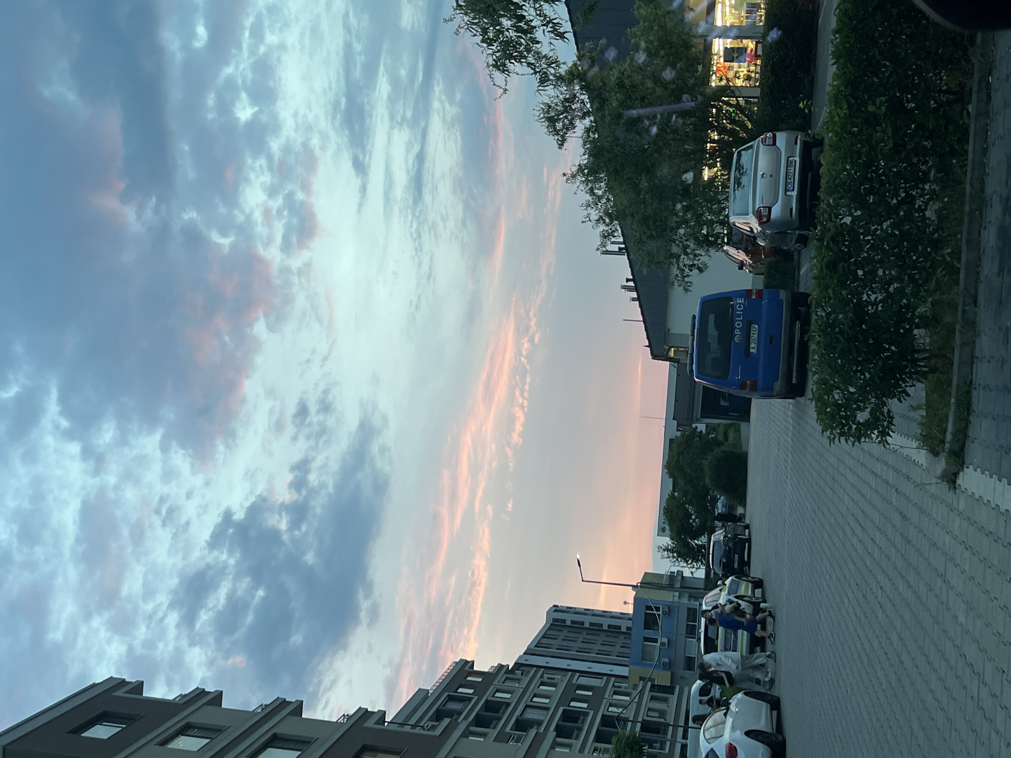 The image depicts an urban street scene at what appears to be sunset or dusk, characterized by a vibrant sky and artificial lights. The sky, occupying a significant portion of the upper frame, transitions from pale blue to soft pink and orange hues, with scattered grey and white clouds.
Along the right side of the image, a paved street or large sidewalk area, constructed from interlocking grey pavers, stretches into the distance. Several vehicles are parked along this area. Prominently visible is a blue van with "POLICE" written in white capital letters on its side, bearing a Bulgarian license plate "J 2874 KP". Behind it, a silver Dacia Logan sedan is parked, with a partially visible Bulgarian license plate "CB 4223 BM". Further down the street, other cars, including a dark sedan and a white car, are parked.
On the left side of the scene, a modern multi-story building with numerous windows and a distinct, stepped architectural design is visible. In the mid-ground, behind the parked cars, another residential or commercial building complex is present, featuring different sections and visible air conditioning units. Further to the right, a commercial building with brightly illuminated windows and interior displays suggests it is open for business.
Scattered greenery, including bushes and trees, lines the street and can be seen between the buildings. A few individuals are visible in the mid-ground, near the parked cars. One person in a blue shirt is interacting with others near a white car. The people's expressions are not discernible, and no specific event is taking place beyond casual interaction in an urban environment. A street lamp extends from the mid-right side of the frame towards the sky. The overall atmosphere suggests a quiet evening in a Bulgarian town, given the context provided.
