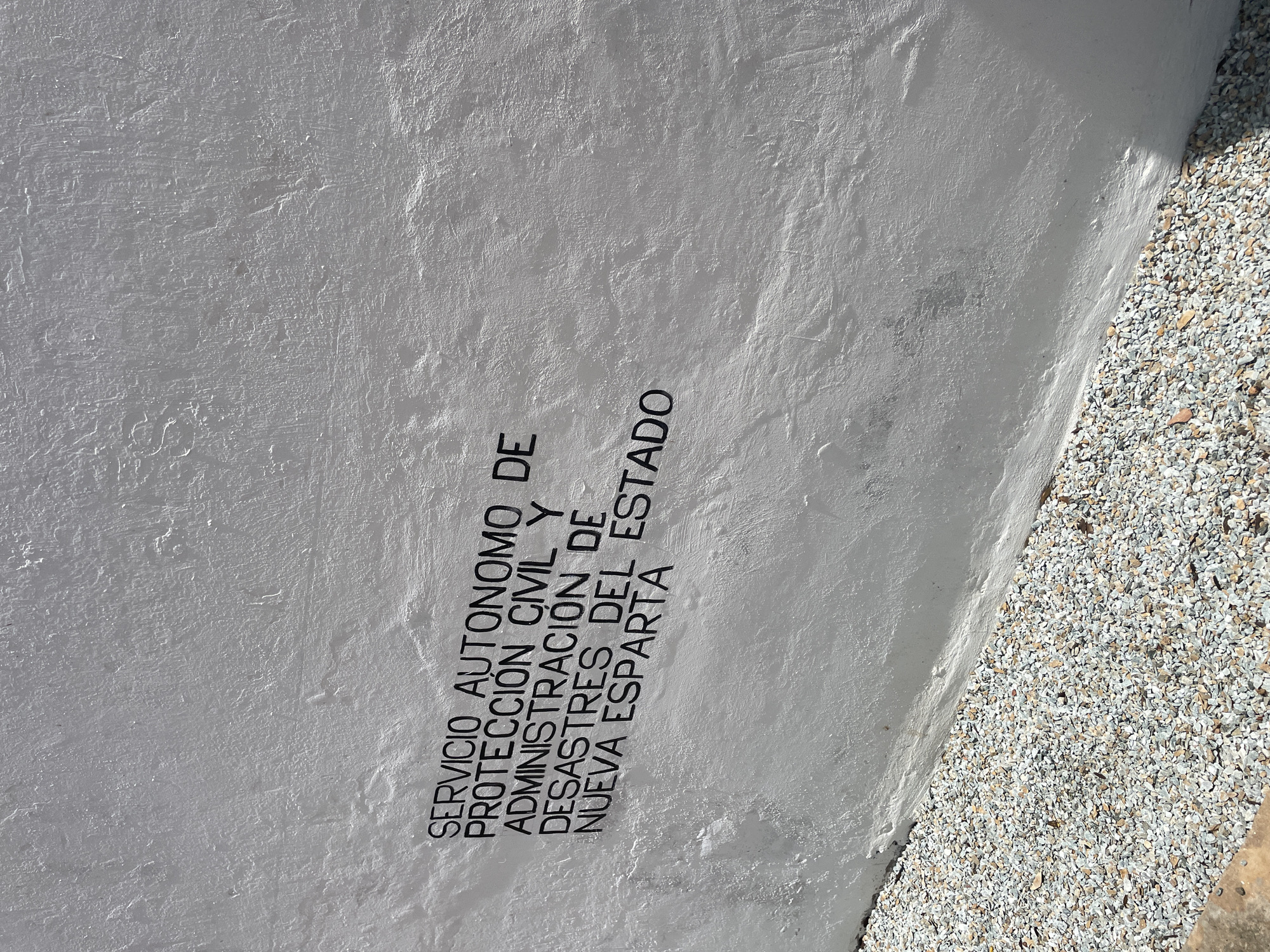 The image shows a section of a rough, white-painted stucco wall with black lettering. The text reads "SERVICIO AUTONOMO DE PROTECCIÓN CIVIL Y ADMINISTRACIÓN DE DESASTRES DEL ESTADO NUEVA ESPARTA." The wall is illuminated by sunlight, casting a shadow on the lower right portion. In the foreground, a band of small, light-colored gravel lines the base of the wall. A small portion of concrete is visible in the bottom left corner. The scene is likely located in El Valle del Espíritu Santo, Venezuela, as indicated by the text on the wall. There are no people or actions depicted in the image.