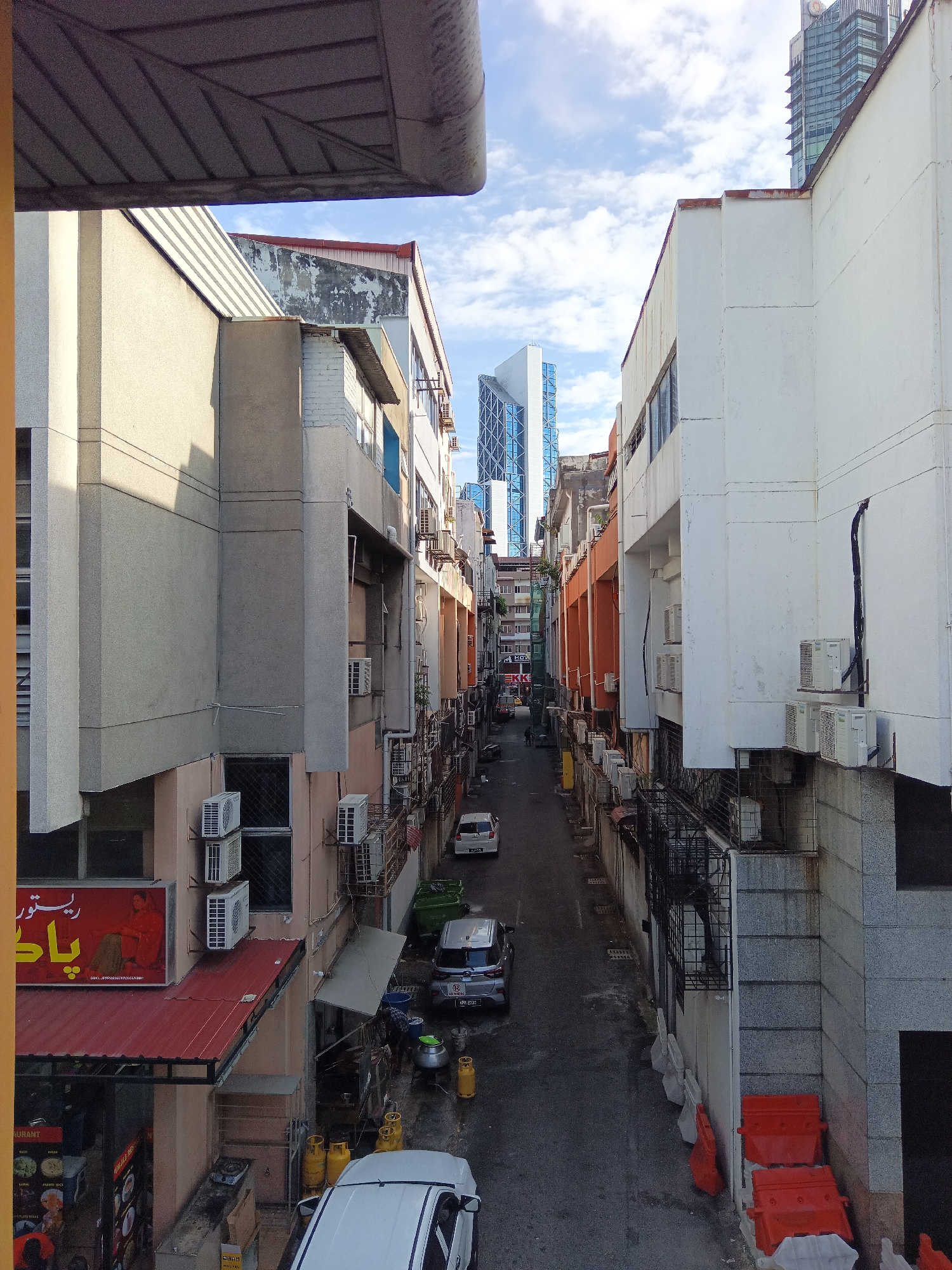 The image captures a narrow street in Kuala Lumpur, Malaysia, lined with multi-story buildings. The architecture suggests a mix of older and more modern structures. Several white air conditioning units are visible mounted on the exterior walls of the buildings. The street is paved and appears to be an alleyway, with parked cars and some orange traffic barriers present. A red-roofed structure, possibly a restaurant with signage, is located on the left side of the image. In the background, taller, modern skyscrapers are visible, indicating the urban setting. The sky is partly cloudy.