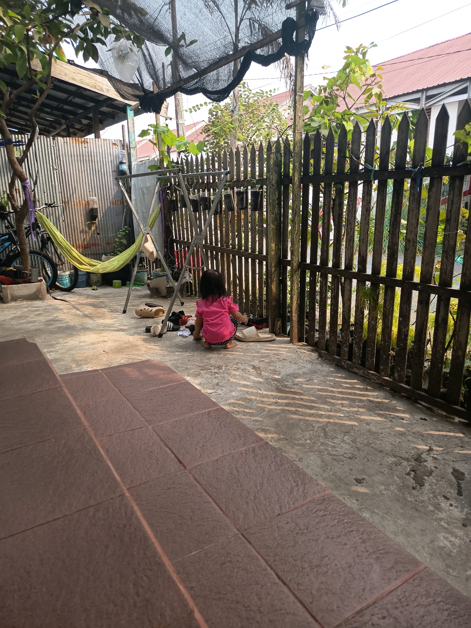 The image depicts an outdoor area with a concrete floor, partially covered by brown tiles in the foreground. A young child, seen from behind and wearing a pink shirt and polka-dot pants, is crouching on the concrete. Next to the child are several items of footwear, including a pair of cream-colored sandals, black and white sneakers, and other dark-colored shoes. In the background, a wooden fence with vertical planks runs along the right side of the frame, with a tree and foliage visible beyond it. To the left, a corrugated metal wall stands near a bicycle, a green hammock suspended from a metal frame, and several buckets. The scene is illuminated by sunlight casting shadows on the ground. This appears to be a residential backyard, potentially located in Banjarmasin, Indonesia, given the architectural style and vegetation.