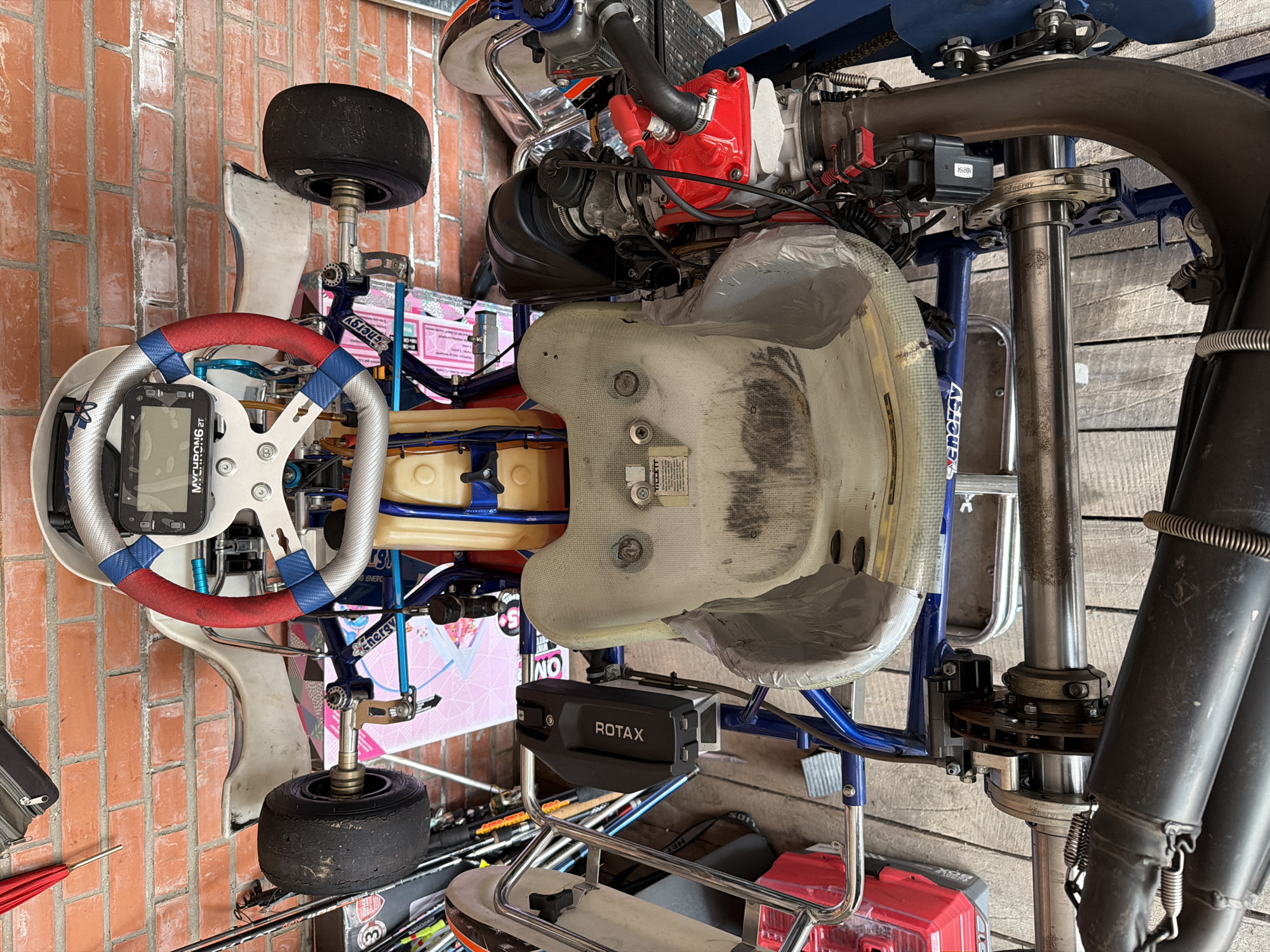 The image shows a go-kart parked indoors, likely in a garage or workshop, with a brick wall and wooden flooring visible in the background. The go-kart is viewed from above and is the central subject. It is white and blue with red and blue accents on the steering wheel. The steering wheel has a digital display. The seat is white and appears worn. The go-kart has a Rotax engine, visible on the right side of the image, which is red and black. The rear wheels and exhaust system are also visible. The overall scene is static, with no people present or any apparent activity. The environment suggests a utilitarian space, possibly associated with racing or go-kart maintenance. The image does not contain any visual cues that would indicate its location in Sukhaya, Russia.