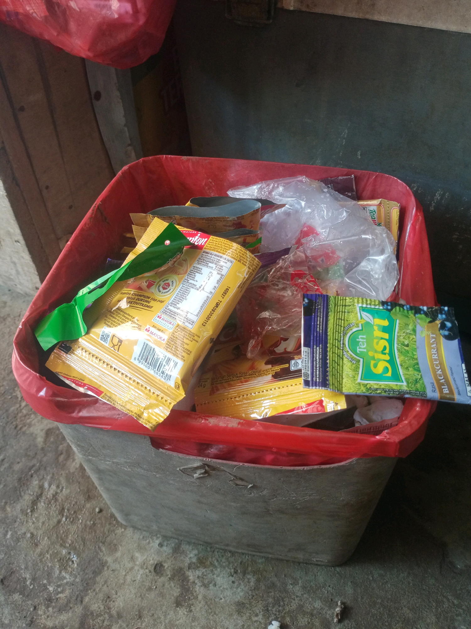 In an outdoor, low-light environment, likely an alley or loading dock, a red plastic trash bin is filled with various discarded items. The bin itself appears weathered and possibly made of concrete or a similar rough material, with a reddish-orange plastic liner visible around the rim. Among the trash, several brightly colored wrappers are prominent, including a yellow packet with nutritional information and Indonesian text, and a green and blue packet labeled "Teh Sisri Blackcurrant." Clear plastic bags, some containing what appear to be food items or wrappers, are also visible. The background shows a rough, wooden wall and a dark, textured surface that might be a building wall or a tarp. The ground beneath the bin is a concrete surface with visible dirt and debris. The image quality is somewhat grainy, suggesting it was taken with a mobile phone camera in low light conditions.