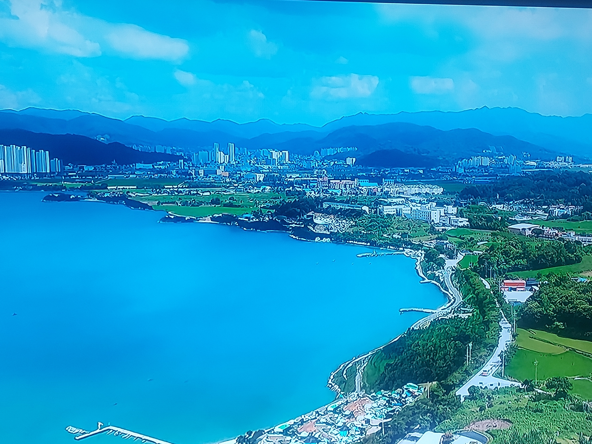 This aerial photograph presents a comprehensive view of a coastal city, identified as Figueira da Foz, Portugal, under bright daylight conditions. A vast expanse of clear, blue-turquoise ocean fills the foreground and left portion of the image, curving along an irregular coastline.
The land features a blend of urban development and natural green spaces. Numerous high-rise buildings, likely residential or commercial, are clustered along the coast and extend inland, forming the city's skyline. These urban areas are interspersed with large sections of lush green, indicating fields, golf courses, or parks, and dense patches of forest. A prominent winding road can be seen tracing the coastline, particularly on the right side of the image. At least two small piers or jetties extend into the water from the shore.
In the far background, a range of dark blue mountains forms a majestic backdrop to the coastal plain and city. The sky overhead is a clear light blue with scattered white clouds, suggesting pleasant weather. The scene captures the harmonious coexistence of a developed city with significant natural elements, including the ocean, mountains, and green landscapes. No specific human activity or visible text can be discerned in this wide, elevated shot.