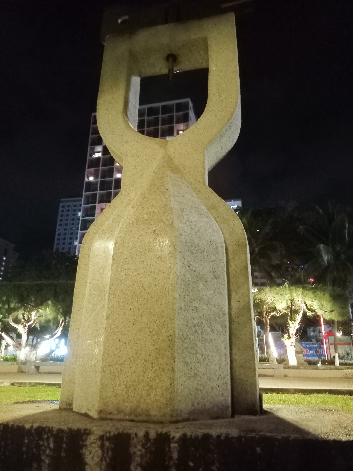 Captured at night, the image focuses on a light-colored, grainy stone sculpture that stands prominently in the foreground. The sculpture has a unique shape, featuring a broader base that tapers inward, then flares out again towards the top into two curved arms that join to form a U-shape. The background reveals elements of an urban environment. A tall, modern building with a grid-like facade is visible behind the sculpture. Trees line the area behind the sculpture, some illuminated by lights that cast a soft glow on their leaves and trunks. Further in the distance, there are glimpses of city lights and buildings, indicating an active urban area. The ground around the sculpture is covered in grass, suggesting a park or public space. Based on the information provided, this scene is located in Nha Trang Ward, Vietnam.