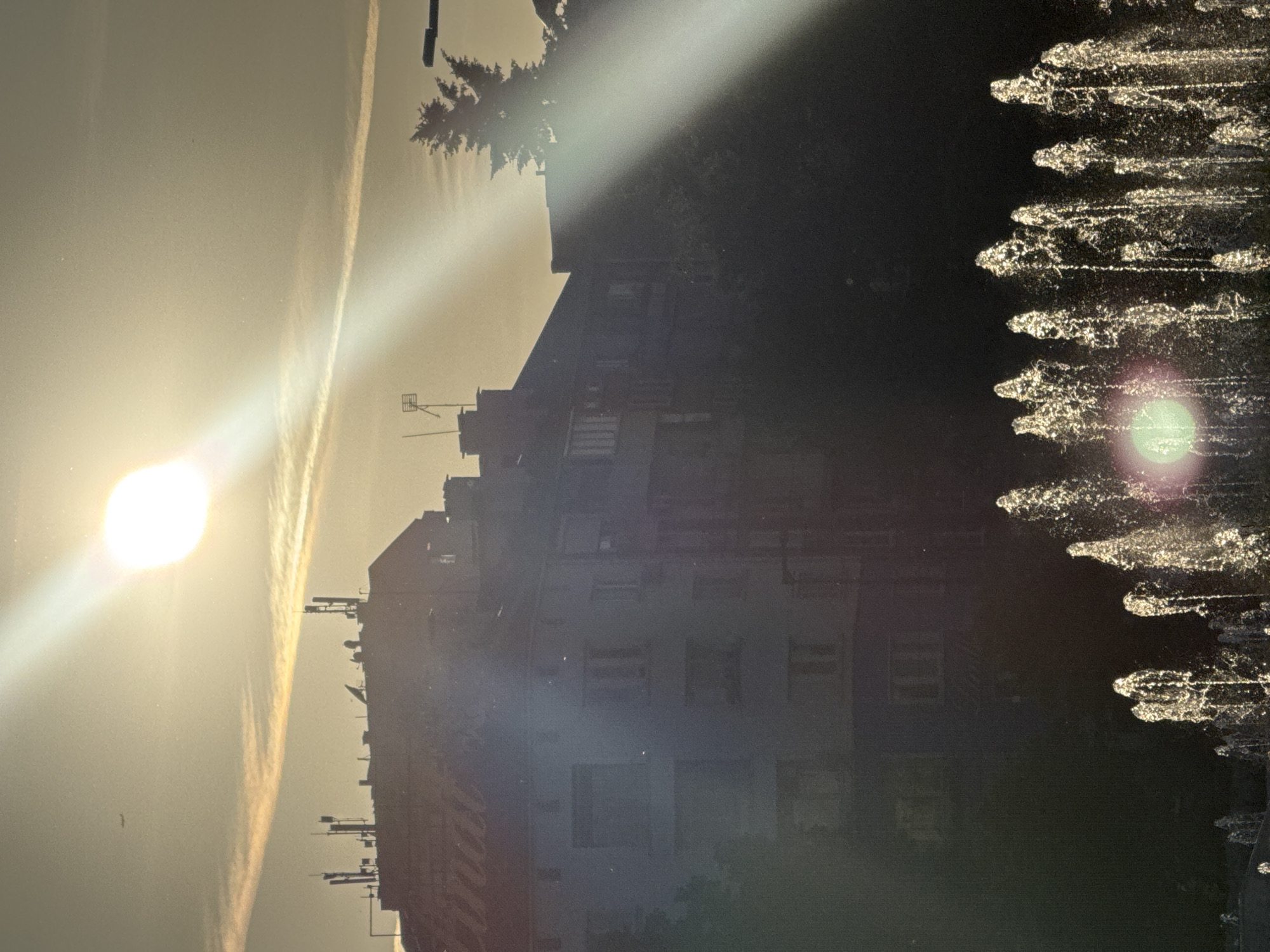 The image shows a building in Sofia, Bulgaria, illuminated by the sun. The building is a multi-story structure with visible windows and architectural details. To the right of the building, a fountain is spraying water. The sky is bright with a contrail.