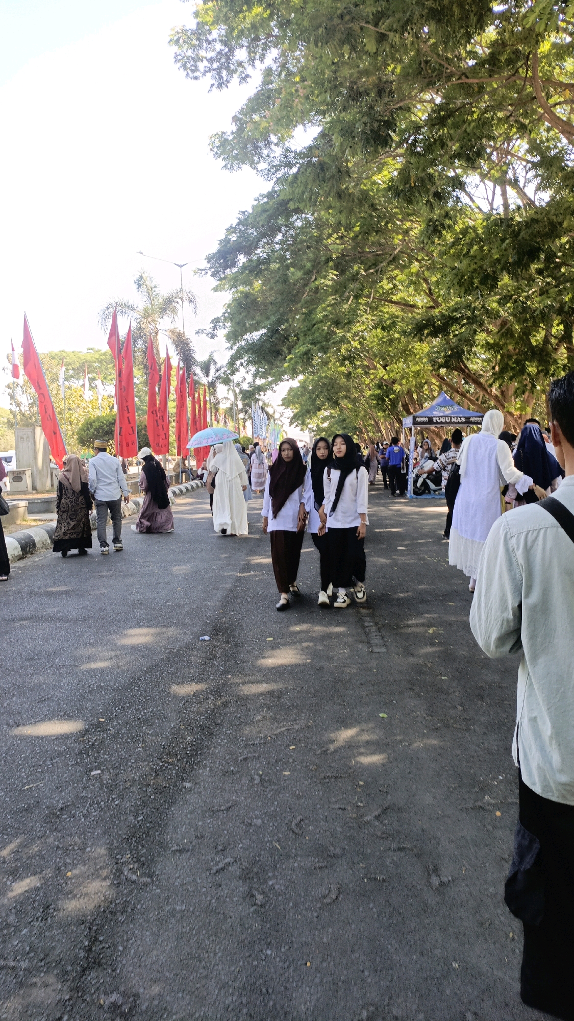 The image captures a street scene in Taliwang, Indonesia, likely during a procession or event. A road paved with asphalt dominates the foreground, with sunlight casting dappled shadows from overhead trees onto its surface. Several people, mostly women wearing hijabs, are walking along the road. Some are in groups, while others are more spread out.
To the left, a row of tall red flags with black lettering is planted along the roadside. In the background, there are trees and some buildings. On the right side of the road, a blue tent with "TUGUMAS" written on it is visible. Behind the tent, more people and some vehicles, including a motorcycle, are present. The overall atmosphere appears to be one of a public gathering or festival.