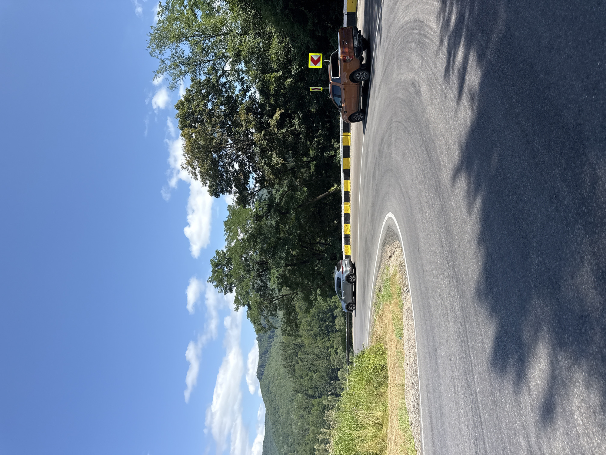 The image depicts a winding asphalt road cutting through a lush, green mountainous landscape under a bright blue sky with scattered white clouds. The road curves sharply, bordered by yellow and black striped barriers. On the right side of the road, a dark orange pickup truck is in motion, with a directional arrow sign indicating a curve ahead. On the left side, a silver SUV is also in motion, navigating the same curve. The surrounding environment is characterized by dense forests covering the hills, with patches of dry grass and vegetation along the roadside. The scene appears to be a scenic route in the Oituz area of Romania, likely a rural or mountainous region.