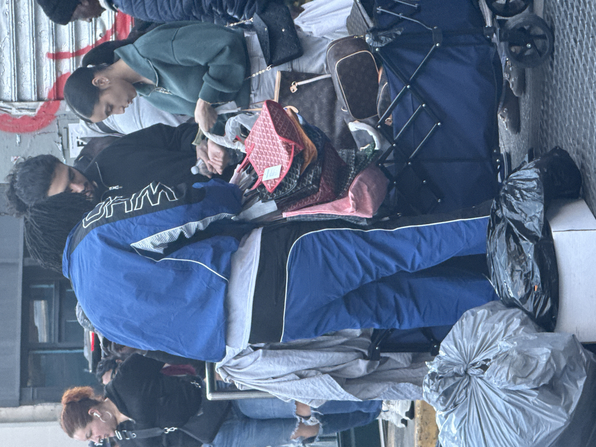 A group of people stand outdoors on a sidewalk. The scene is cluttered with items, including bags and what appears to be a foldable wagon. The individuals are clad in various casual clothing. The location is implied to be within New York City, USA.