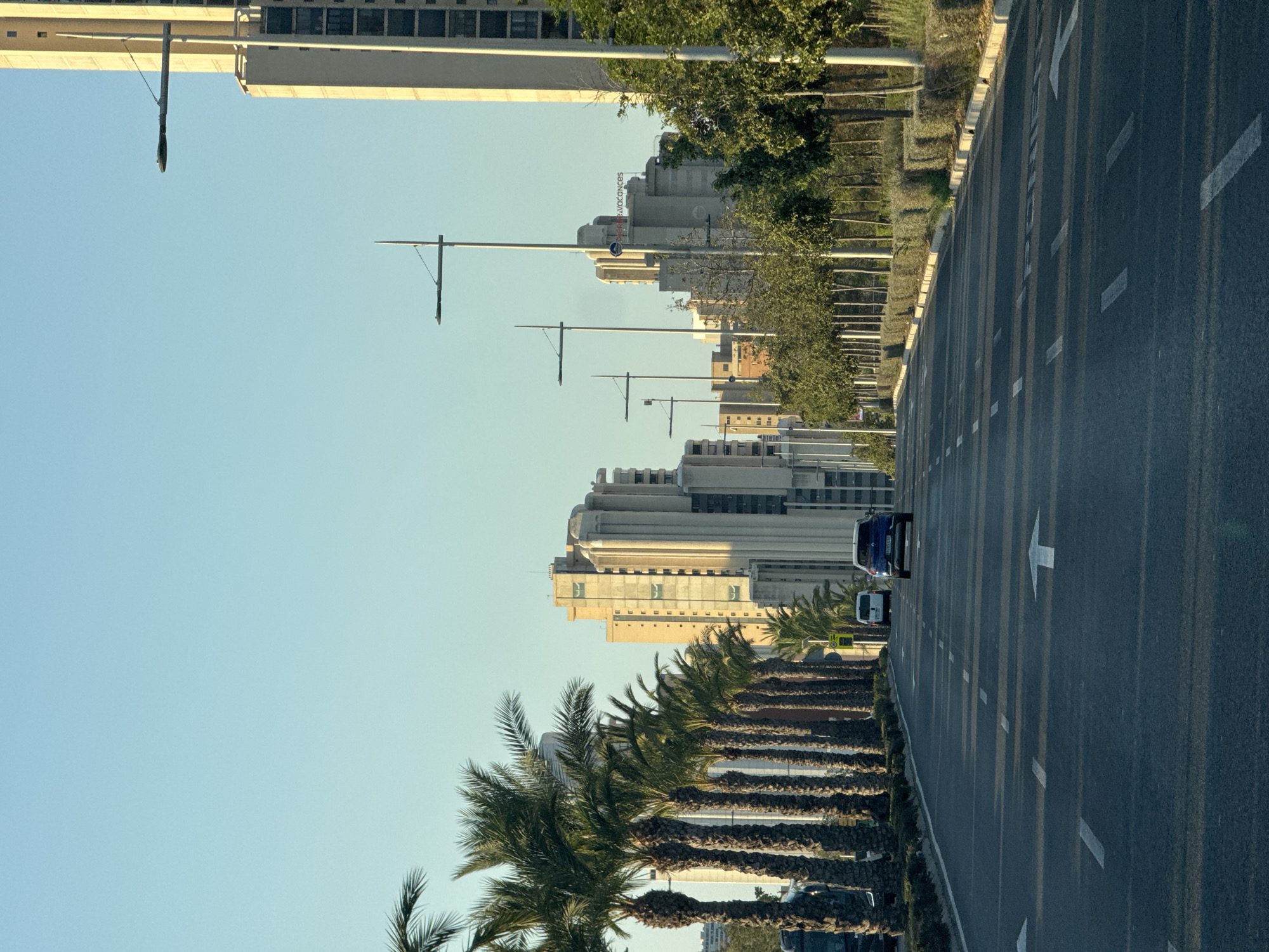 The image shows a view down a wide, multi-lane asphalt road in Benidorm, Spain. Tall buildings, characterized by their art deco style and light-colored facades, line the right side of the road, interspersed with trees. A row of mature palm trees stands on the left side, casting long shadows across the road. The sky is clear and blue.
Several vehicles are visible on the road: a blue car in the foreground, a white van further ahead, and another vehicle partially obscured by the palm trees on the left. Streetlights with a modern, sleek design are positioned along the roadside, some attached to tall poles and others extending from the sides of the buildings.
The road has clear white lane markings and directional arrows painted on its surface. A low, manicured hedge runs along the side of the road, separating the pavement from the buildings and trees. The overall impression is one of a modern, well-maintained urban environment, likely a major thoroughfare in the city. The angle of the shadows suggests the photograph was taken in the late afternoon or early morning.
