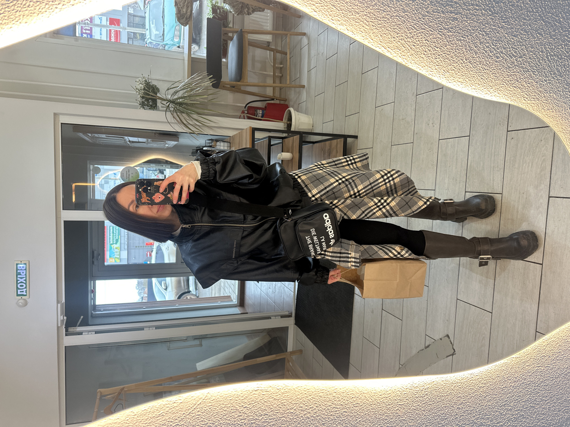 A person is reflected in a mirror, taking a picture with a smartphone. The person is wearing a black jacket, a gray plaid skirt, black leggings, and brown boots. They are also carrying a black Adidas shoulder bag. The location appears to be inside a building, possibly a retail space, in Votkinsk, Russia. The reflections visible through the door and windows show a city scene with vehicles and buildings. A sign above the door reads "EXIT" in Russian.