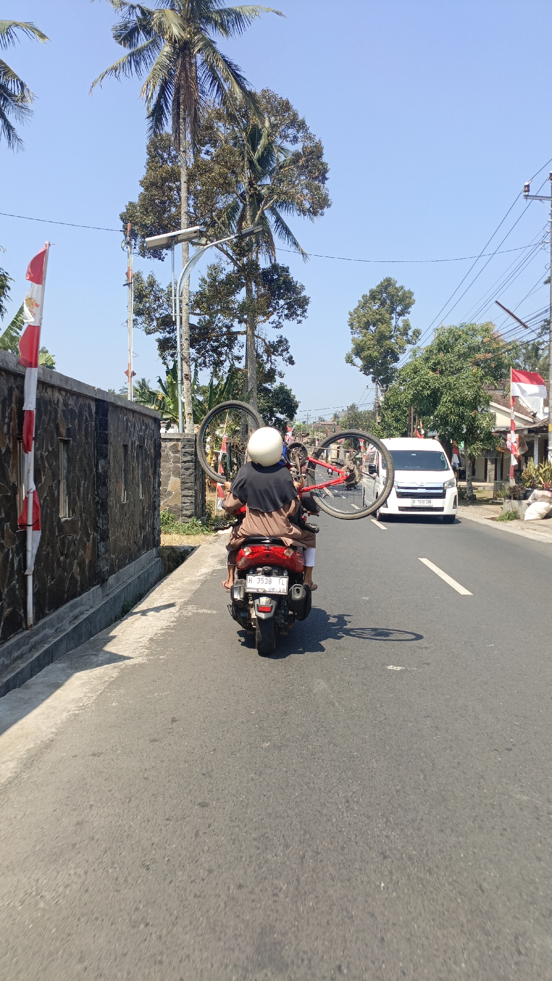 In Ujung-ujung, Indonesia, two individuals are riding on a red scooter along a paved road on a sunny day. The person in the back, wearing a white helmet and a brown outfit with a dark shawl, is carrying a disassembled red bicycle over their shoulder. The scooter has a license plate that reads "H 3538 TC". A white van is visible on the road ahead of them, and to the left, a stone wall runs alongside the road. Tall trees, including a coconut palm, and electrical poles are present in the background. Indonesian flags, red and white, are also visible along the roadside. The road is marked with white lines, and shadows from the scooter and trees are cast on the asphalt.