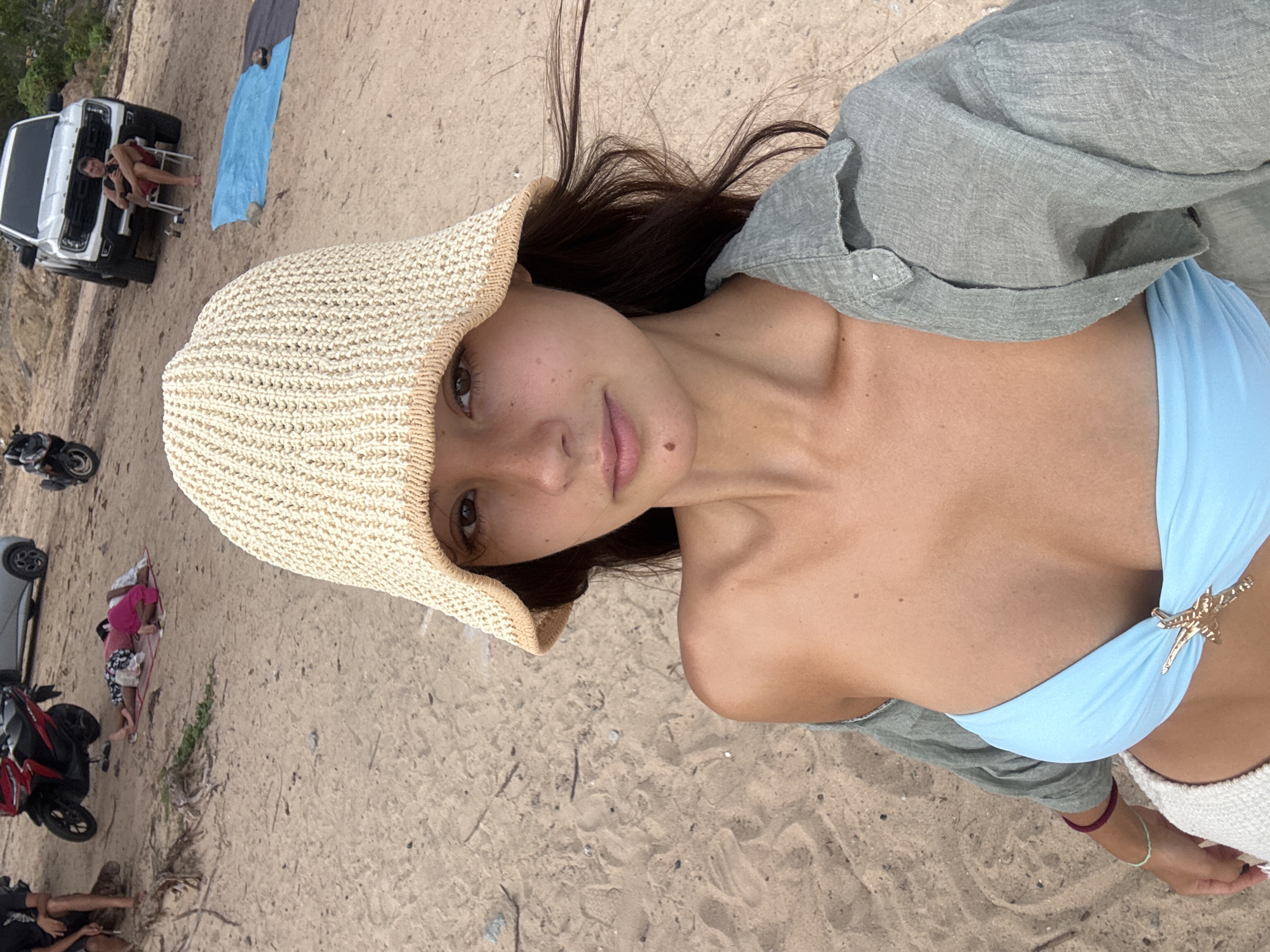 A woman wearing a light blue bikini top, a light green linen shirt, and a straw hat is lying on a sandy beach. The sand appears to be light in color. In the background, there is a white truck parked on the sand, with a person sitting in a chair next to it. Nearby, there are motorbikes and people on the beach. The image appears to be taken on a beach in Rawai, Thailand.