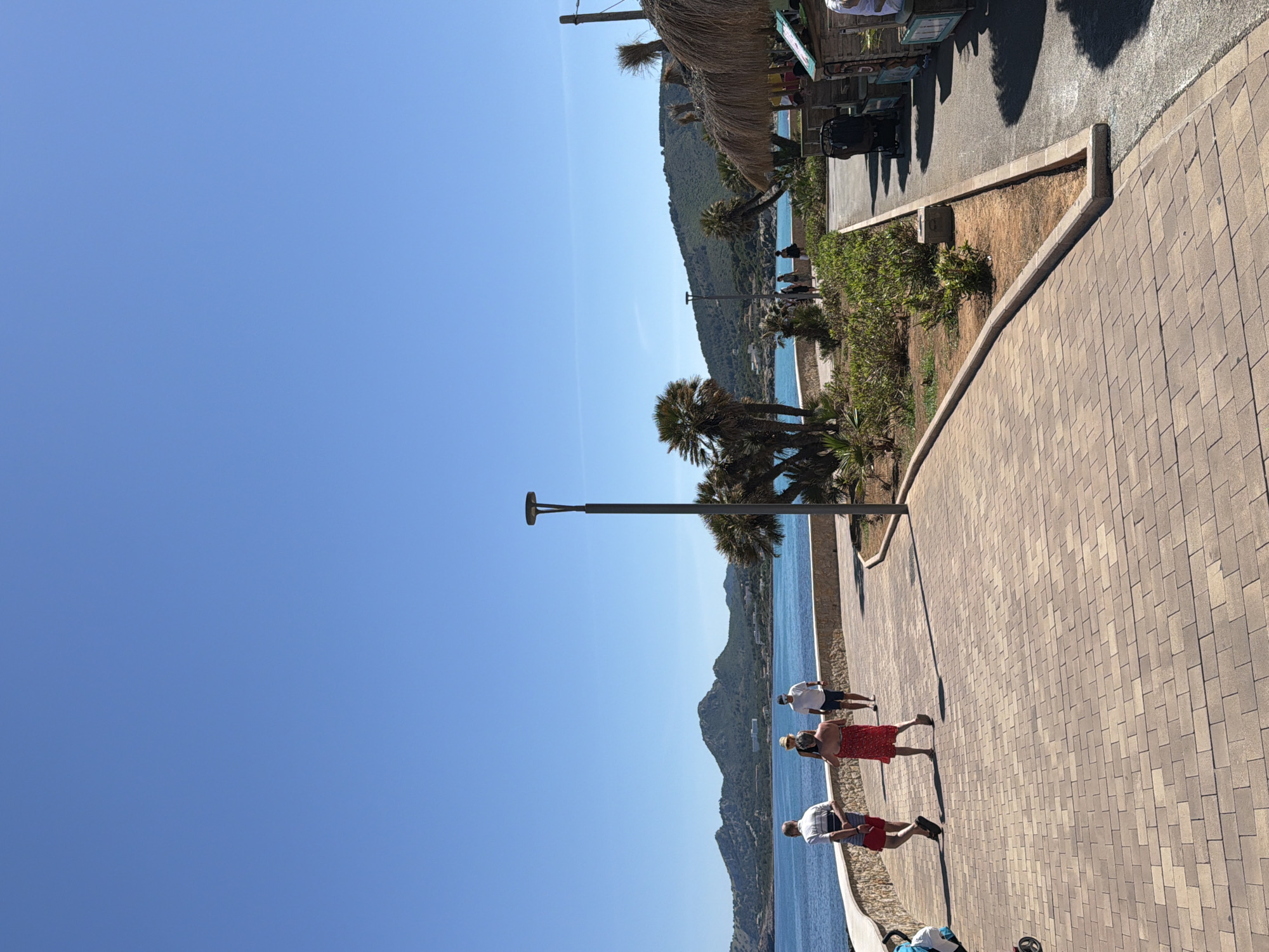 The image captures a coastal scene in Capdepera, Spain, under a clear, bright blue sky. The vantage point appears to be along a paved walkway made of interlocking brick-like stones.
The foreground is dominated by the walkway. To the right of the walkway is an area of shrubbery and then a narrower paved surface.
Several people are seen walking along the walkway, including two people walking side by side, one wearing a red dress and the other a light shirt. Other people are seen further away.
Beyond the walkway and a short vertical drop, there is a stretch of blue water, bordered by land with lush vegetation and low-lying mountains. The landscape forms a coastline.
In the upper right corner of the image, there is what appears to be a structure with a straw or thatched roof, possibly part of a beachside establishment or kiosk.
A tall light pole is visible towards the middle of the image, running vertically, situated close to the edge of the walkway.
