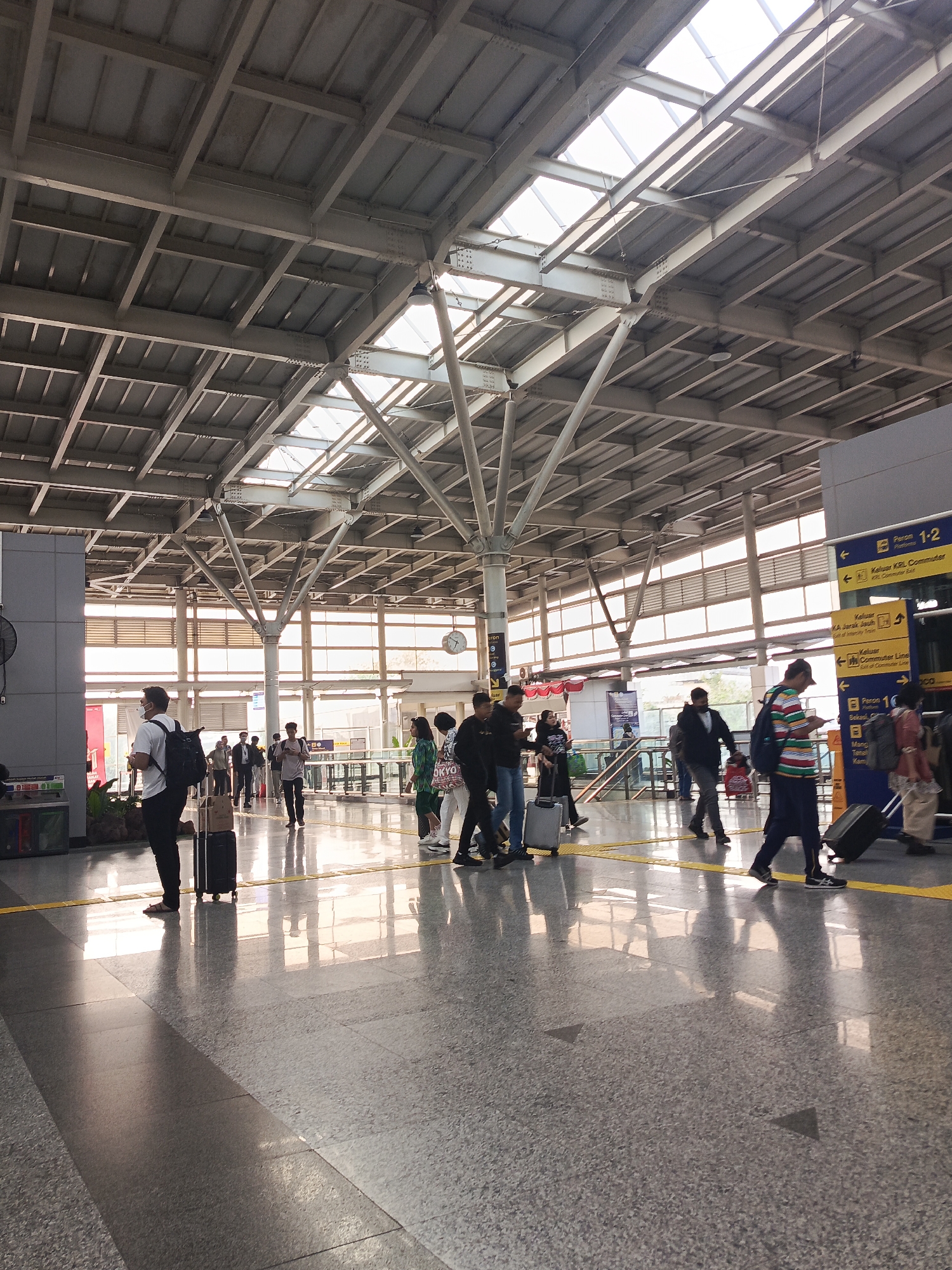 The image depicts the interior of a modern train station, possibly in East Jakarta, Indonesia, with a high, industrial-style ceiling featuring exposed beams and translucent panels that allow natural light to filter through. The floor is polished and reflective, showing the reflections of the people and the ceiling structure.
Several individuals are visible, most of whom are walking and appear to be travelers. Many are carrying luggage, including rolling suitcases and backpacks. Some people are walking alone, while others are in small groups. One person in the foreground is wearing a white t-shirt and black pants, with a white backpack and a black rolling suitcase. Another individual further back is wearing a striped shirt and dark pants, pulling a black rolling suitcase.
Signage is present on the right side of the image, indicating platforms and directions for commuter lines and intercity trains. The overall atmosphere suggests a busy but orderly transit hub. The architecture points towards a functional and contemporary design typical of many transportation facilities.